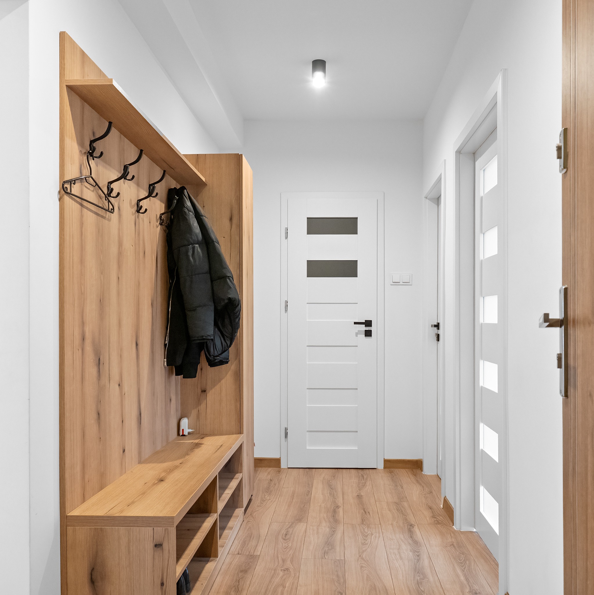 Nowoczesny przedpokój z drewnianym panelem ściennym, wieszakami i siedziskiem. Na wieszaku wisi czarna kurtka. Białe drzwi z przeszkleniami i drewniana podłoga.