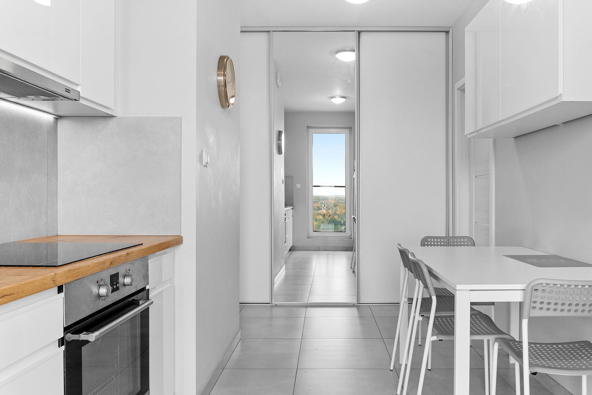 Jasna kuchnia z drewnianym blatem i widokiem na dalszą część mieszkania przez przesuwne drzwi. Minimalistyczny design, dominująca biel, nowoczesne oświetlenie.