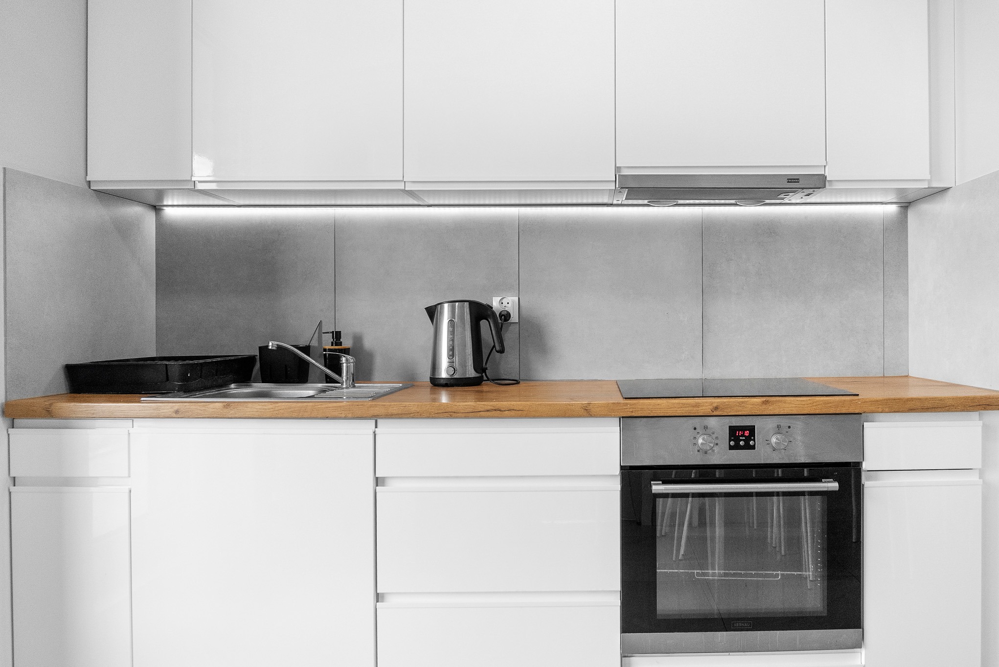 Minimalistyczna, biała kuchnia z drewnianym blatem, srebrnym czajnikiem i piekarnikiem. Podświetlenie LED nad blatem. Nowoczesny design, czyste linie.