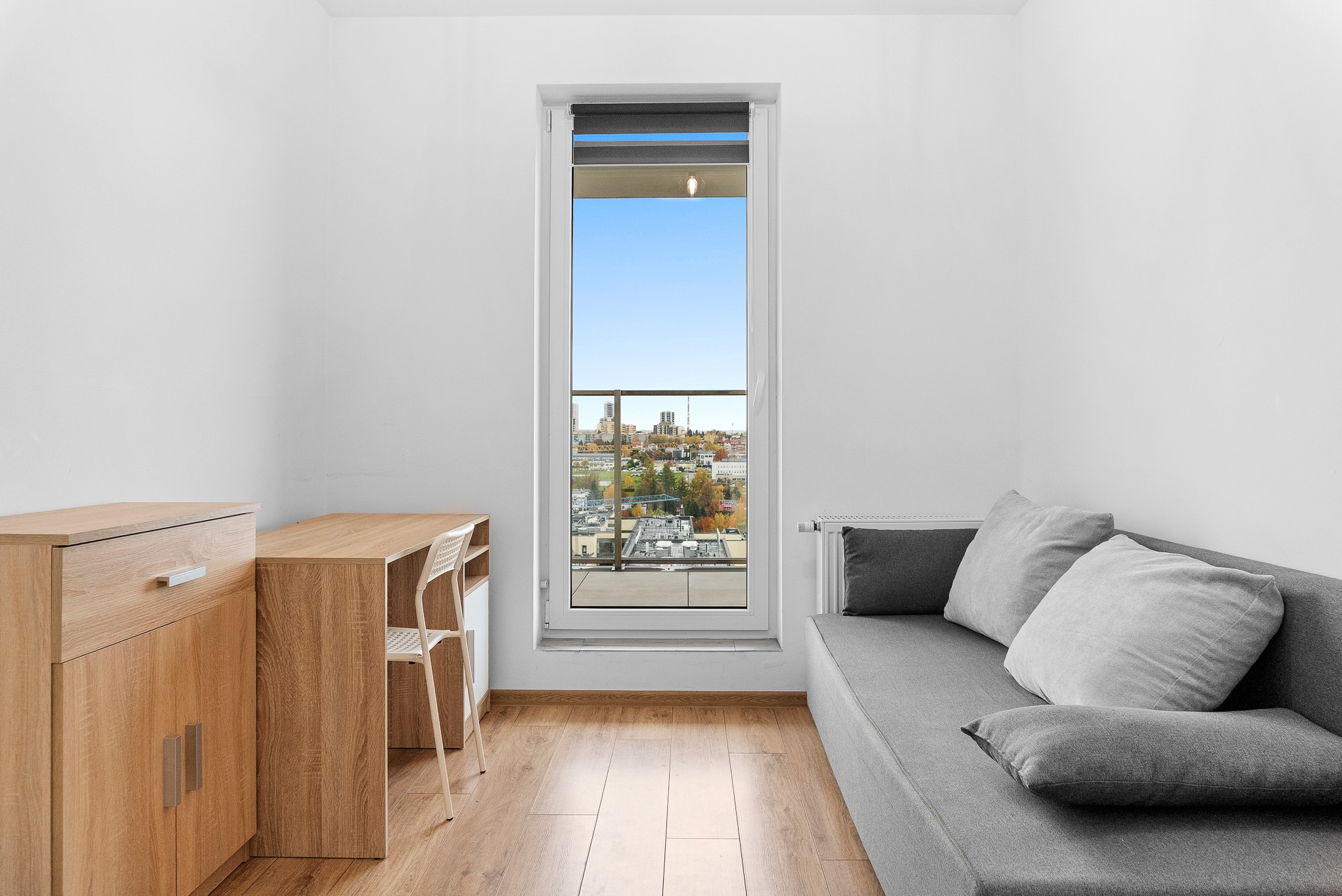 Jasne wnętrze pokoju z biurkiem, szafką i szarą sofą pod oknem z widokiem na miasto. Minimalistyczny design, drewniana podłoga, białe ściany.