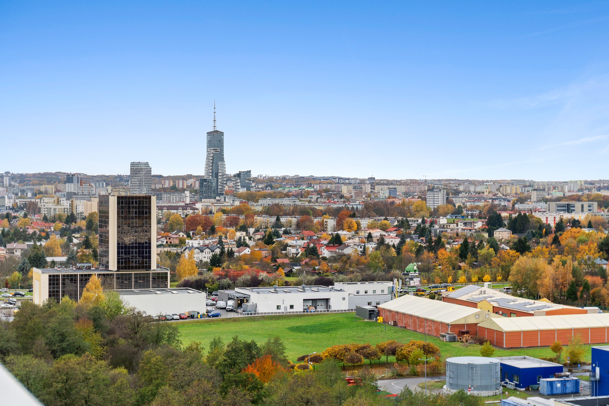 Panorama miasta z wieżowcami w tle, widok z góry na budynki, drzewa w jesiennych barwach i błękitne niebo. Ujęcie z perspektywy.