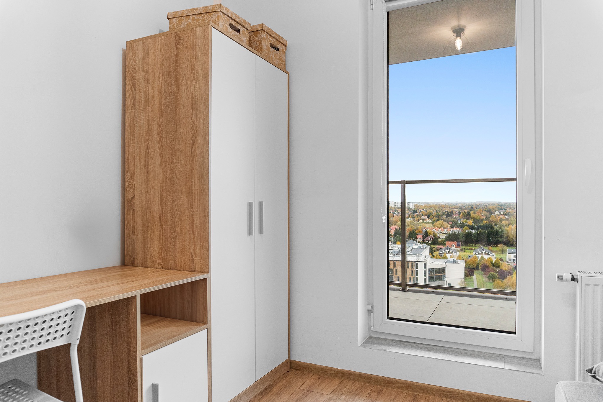 Jasne wnętrze pokoju z biurkiem, szafą i oknem balkonowym z widokiem na miasto. Minimalistyczny design, drewniane akcenty, naturalne światło.