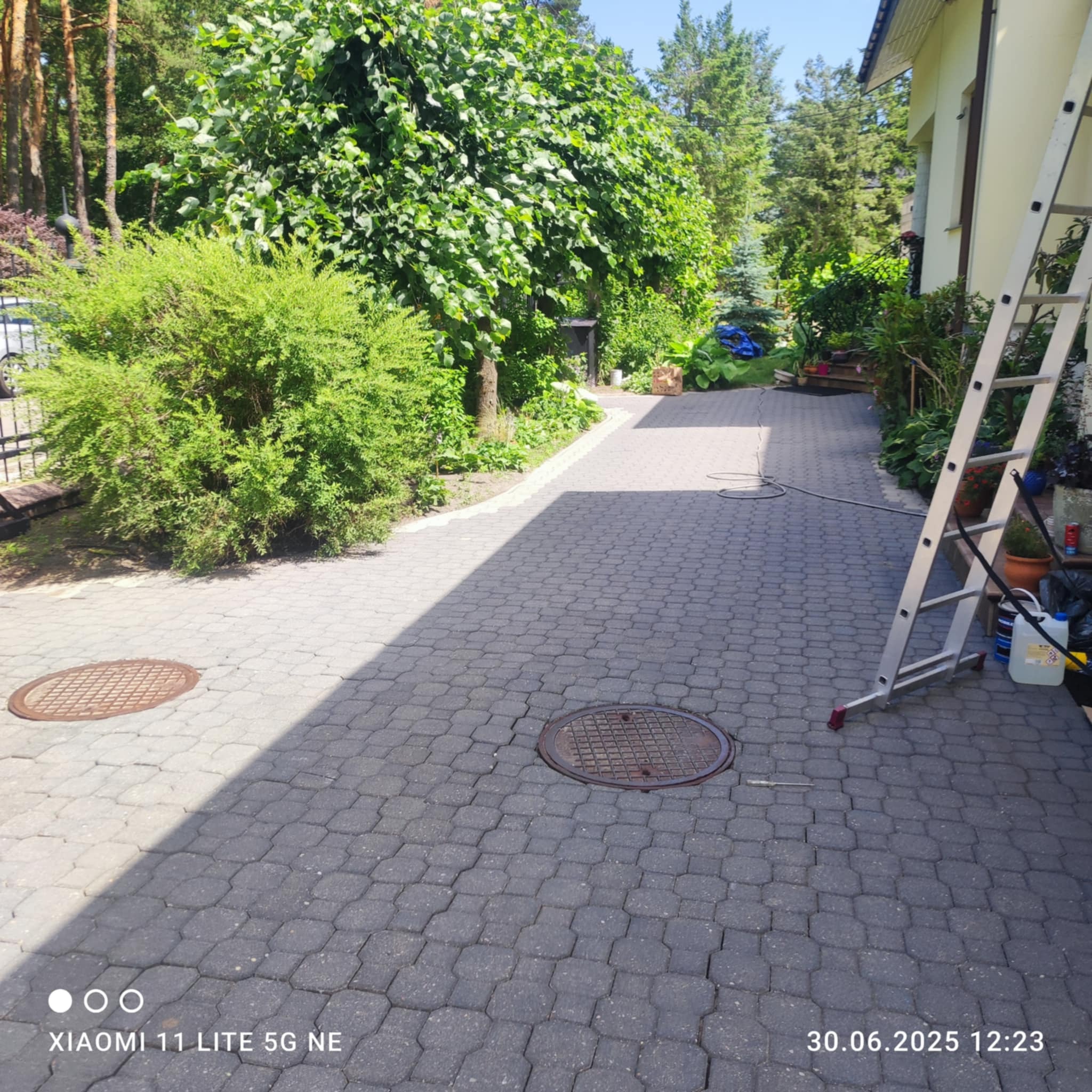 Kostka brukowa przed domem, częściowo w cieniu, z widoczną drabiną i studzienkami. Zieleń w tle. Ujęcie z poziomu gruntu, w słoneczny dzień.