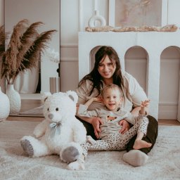 PHOSPHOROS AGNIESZKA RUSINEK - Uśmiechnięta mama z synkiem siedzą na puszystym dywanie obok pluszowego misia. W tle dekoracyjny kominek i wazony z pampasami. Ciepłe, naturalne światło.