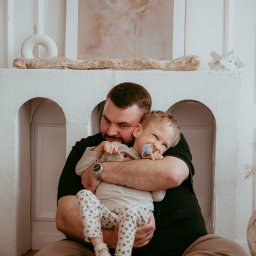 PHOSPHOROS AGNIESZKA RUSINEK - Uśmiechnięty tata tuli synka z smoczkiem na tle białego kominka z dekoracjami. Ciepłe, naturalne światło podkreśla intymną chwilę na sesji rodzinnej.