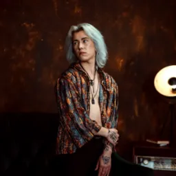 Osoba z platynowymi włosami i tatuażami na dłoniach, w otwartej, wzorzystej koszuli, siedzi na ciemnozielonej sofie na tle brązowej ściany. Obok stoi lampa i radio.