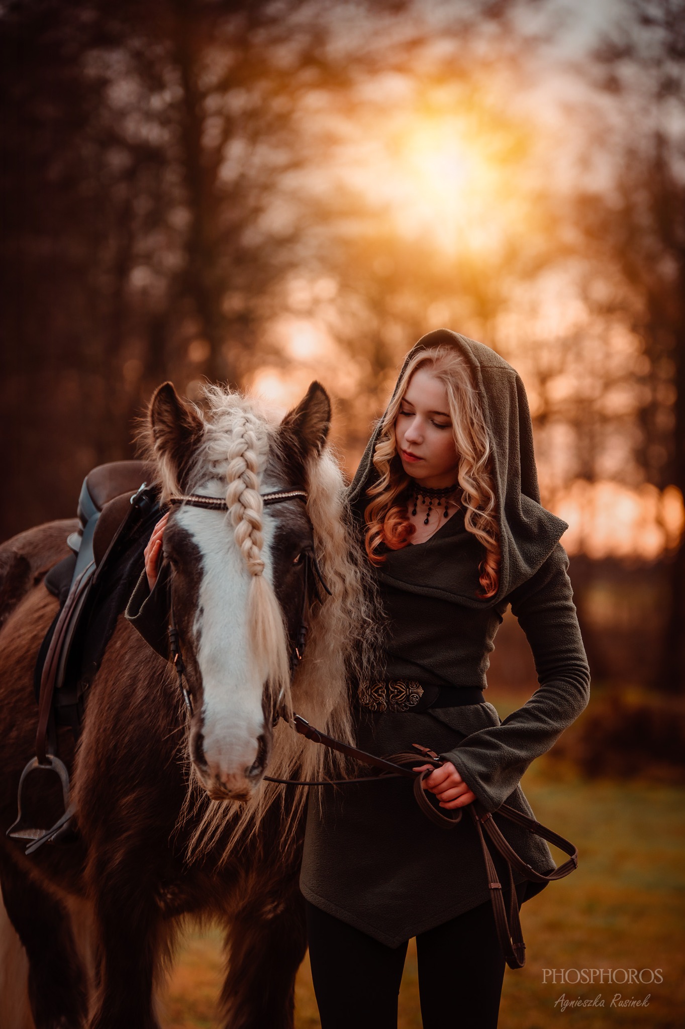 Kobieta w kapturze i stylizowanej sukni pozuje z koniem na tle jesiennego krajobrazu. Uwagę przykuwa zapleciona grzywa konia i ciepłe światło zachodzącego słońca.