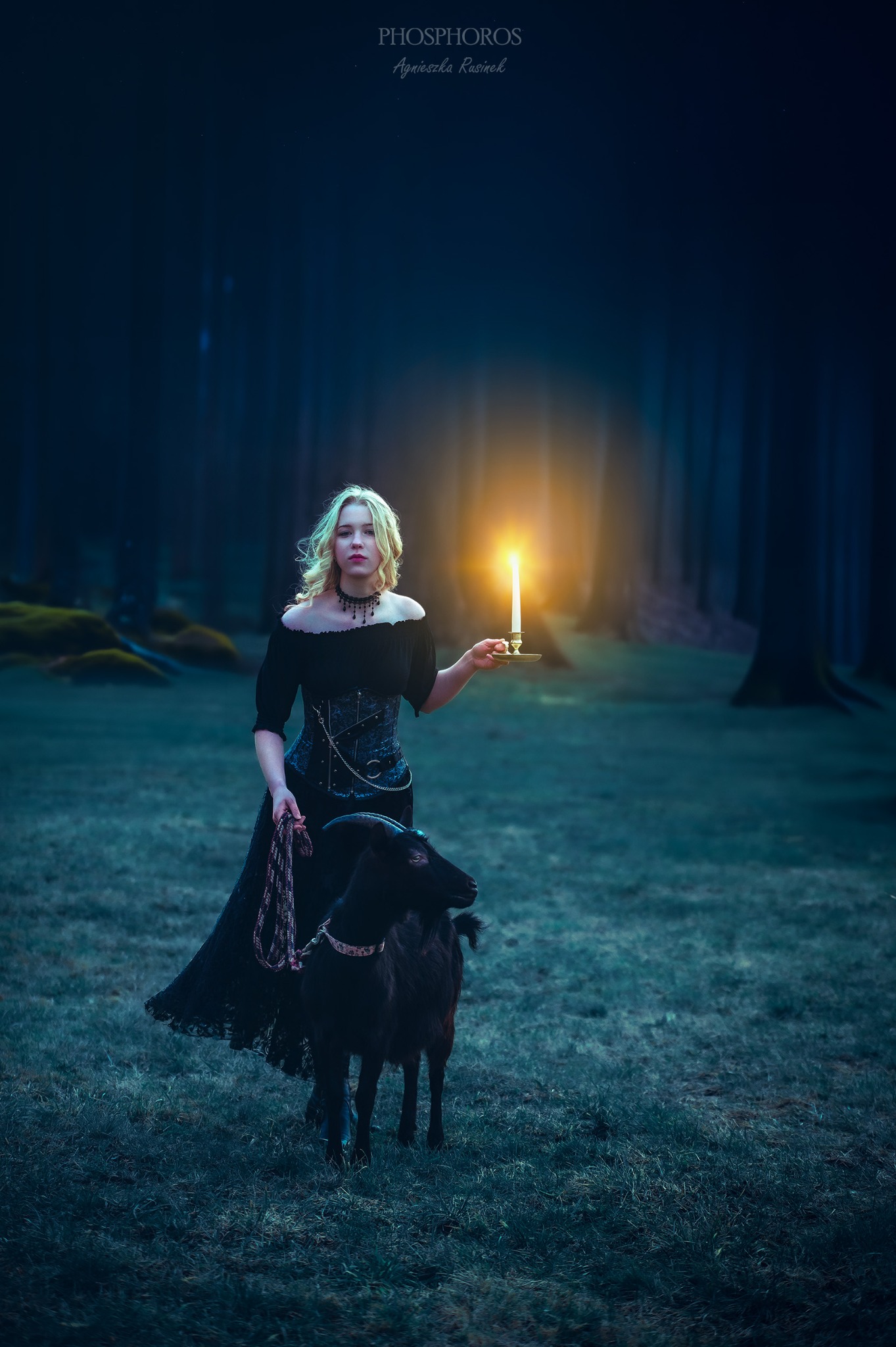 Kobieta w czarnej sukni z kozą na tle ciemnego lasu, trzymająca świecę. Artystyczne ujęcie z delikatnym światłem i mrocznym klimatem. Sesja w plenerze.