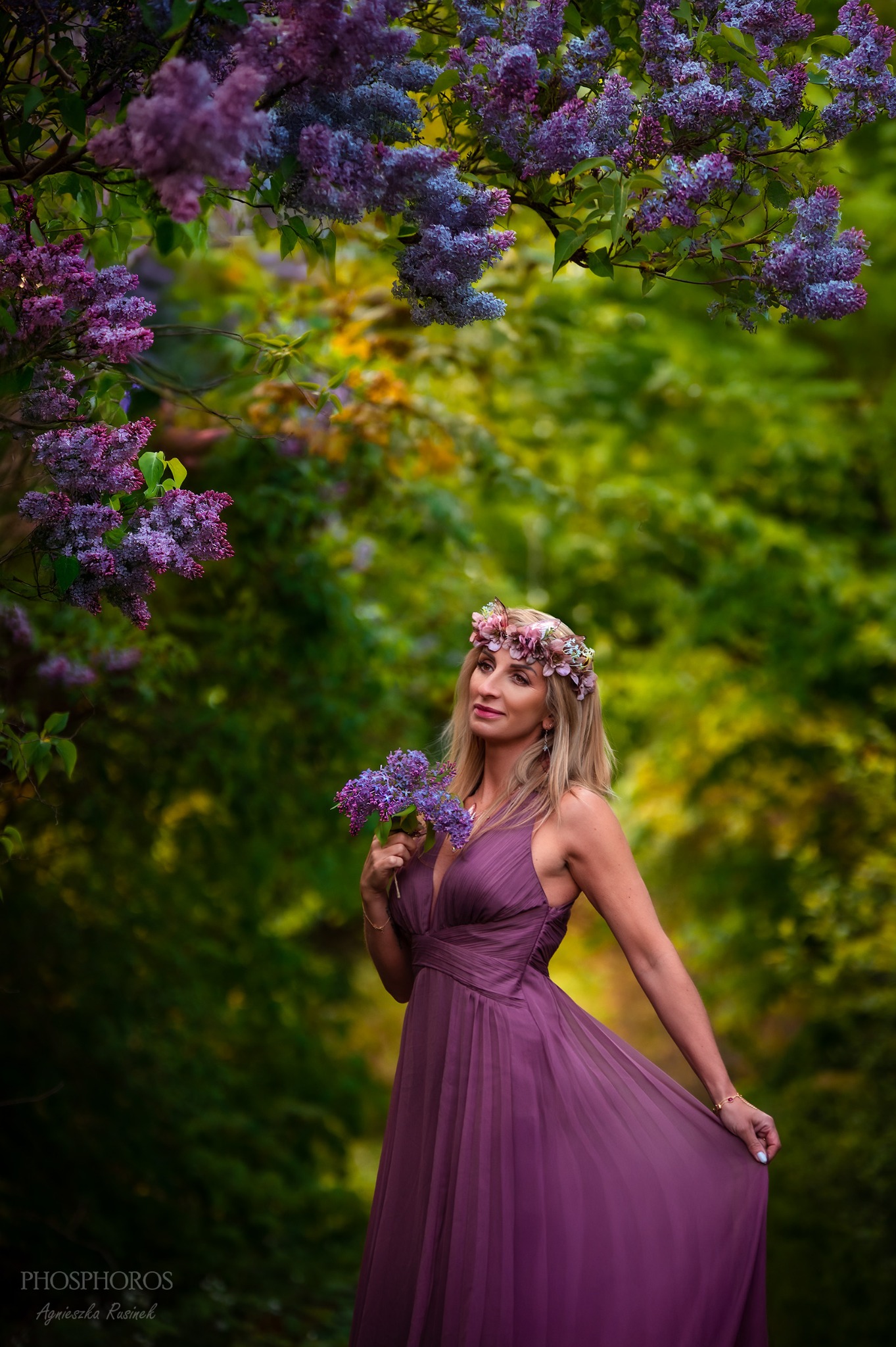 Kobieta w fioletowej sukni i wianku z kwiatów trzyma bukiet bzów na tle zieleni. Portret w naturalnym świetle, z delikatnym rozmyciem tła.