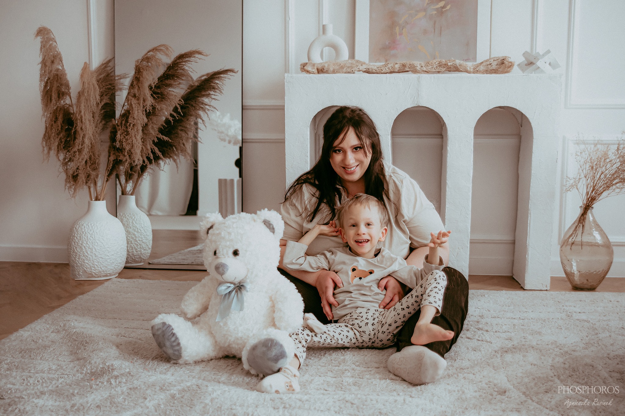 Uśmiechnięta mama z synkiem siedzą na puszystym dywanie obok pluszowego misia. W tle dekoracyjny kominek i wazony z pampasami. Ciepłe, naturalne światło.