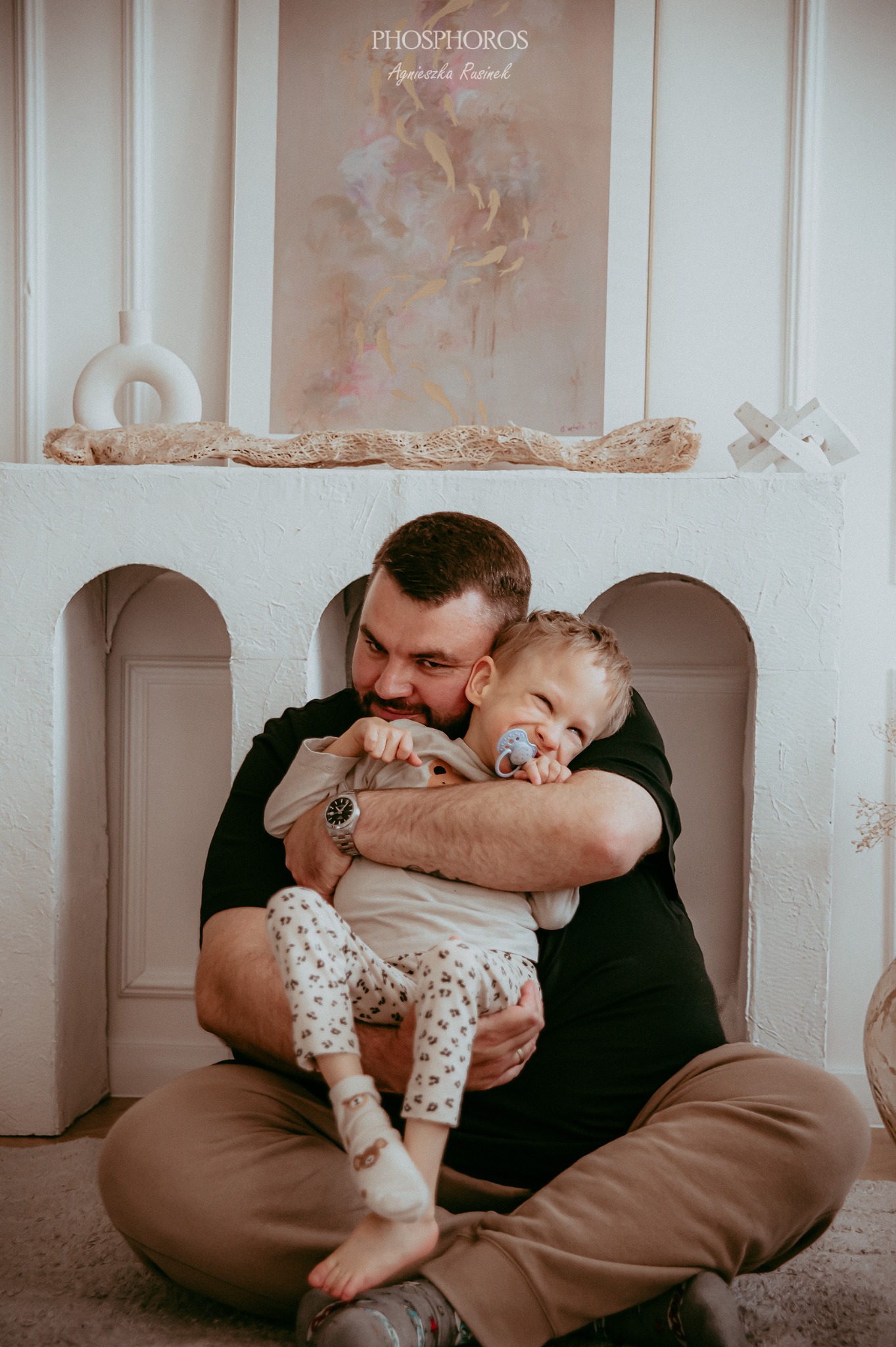 Uśmiechnięty tata tuli synka z smoczkiem na tle białego kominka z dekoracjami. Ciepłe, naturalne światło podkreśla intymną chwilę na sesji rodzinnej.