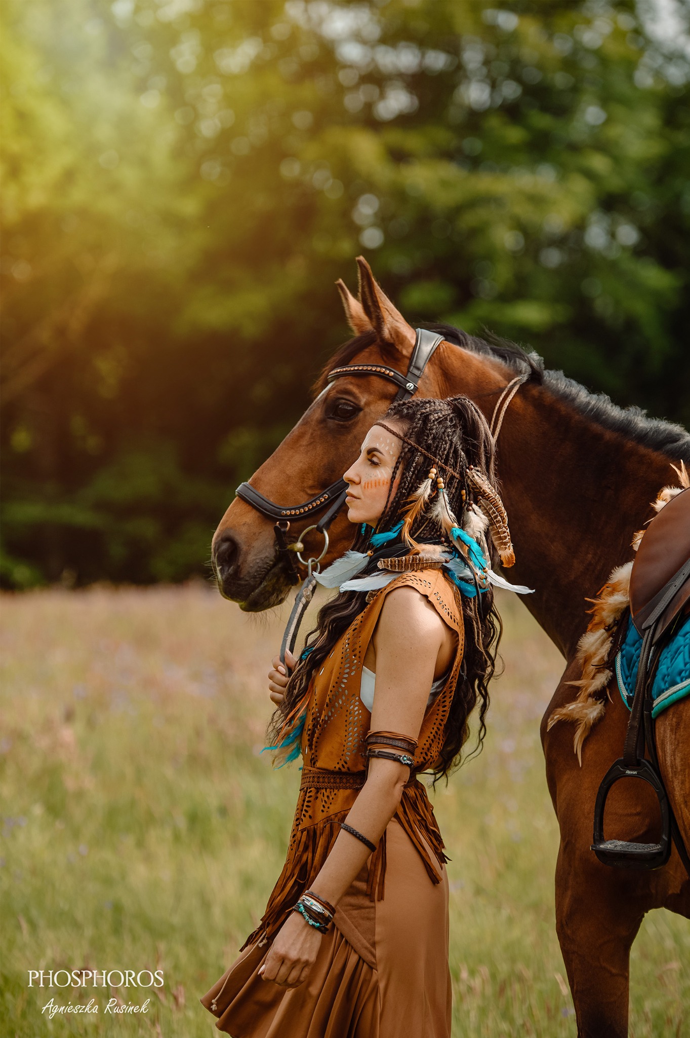 Kobieta w stylizacji indiańskiej z piórami i malunkami na twarzy, pozuje z brązowym koniem na tle zieleni. Ujęcie w plenerze, ciepłe światło.