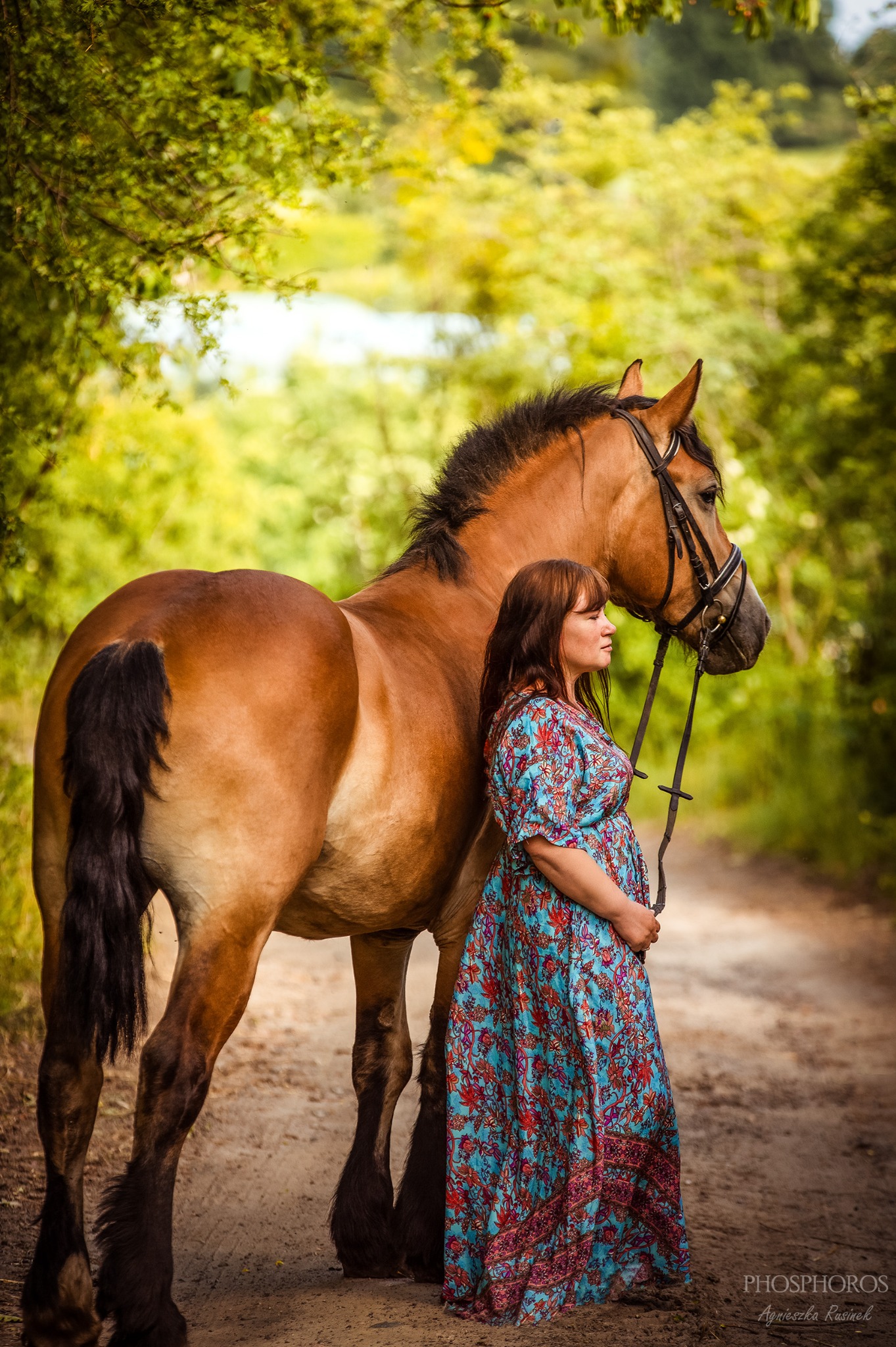 Kobieta w kwiecistej sukience, stojąca obok brązowego konia na leśnej ścieżce. Romantyczna sceneria, miękkie światło, naturalne kolory. Delikatny uśmiech kobiety.
