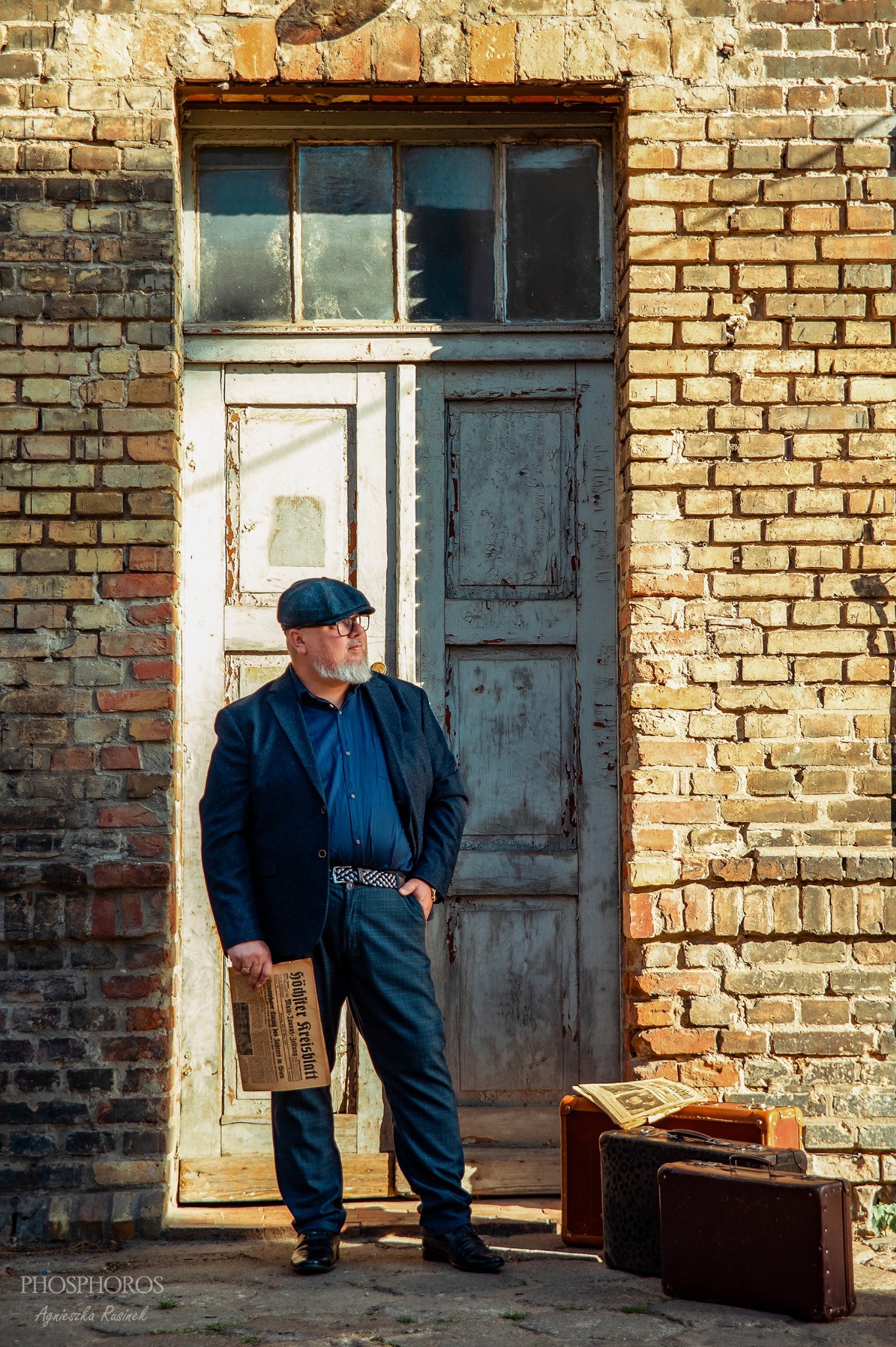 Mężczyzna w garniturze i kaszkiecie, z gazetą w dłoni, stoi przed ceglaną ścianą z drzwiami. Obok leżą walizki. Ujęcie w ciepłych barwach, w stylu vintage.