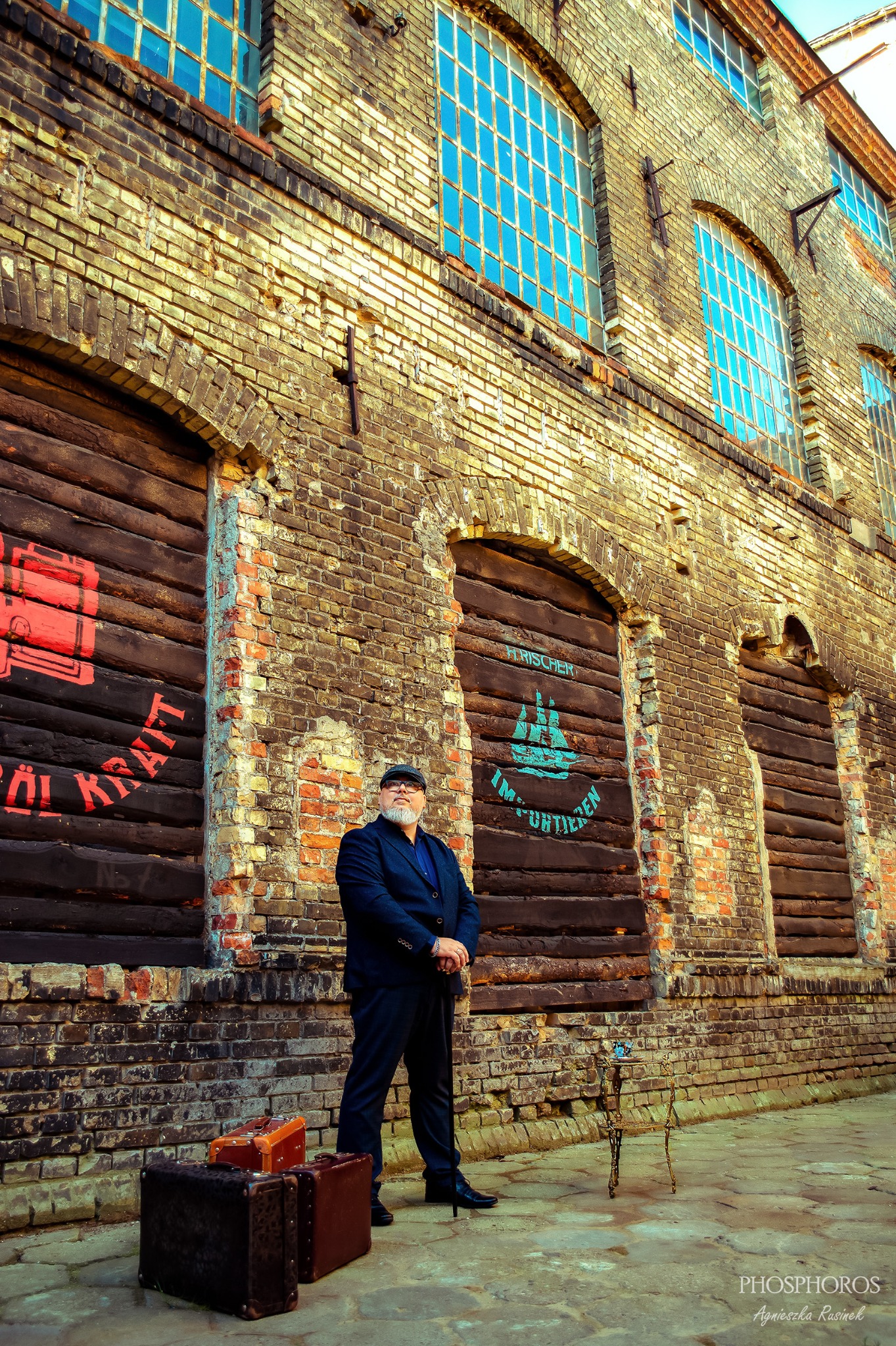 Mężczyzna w garniturze i kaszkiecie pozuje na tle ceglanej ściany z drewnianymi, pomalowanymi drzwiami, obok leżą walizki, a w tle widać okna.