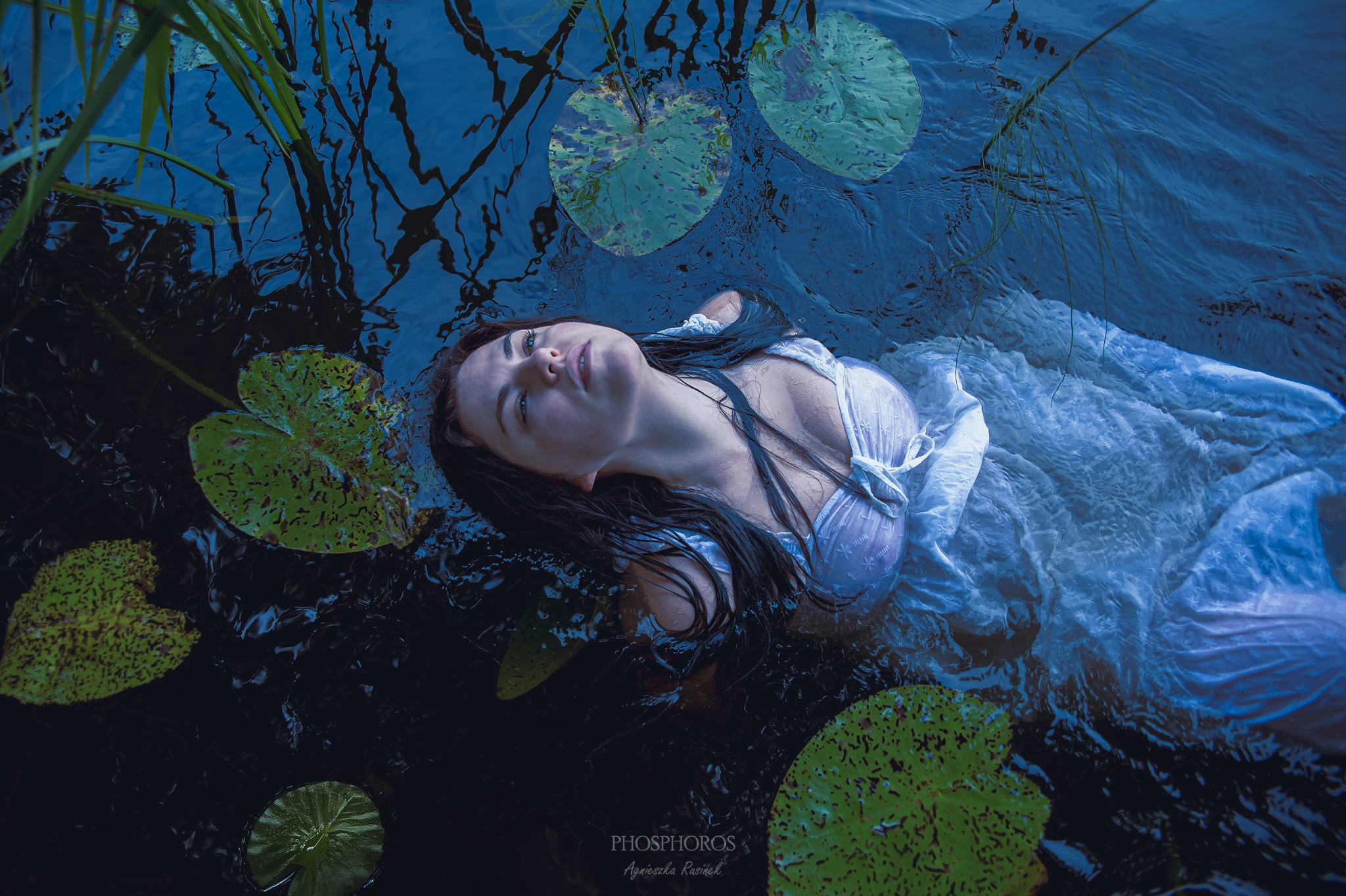 Artystyczna sesja kobieca: brunetka w zwiewnej sukience unosi się na wodzie wśród lilii wodnych, patrząc w górę, w chłodnej, niebieskiej tonacji.