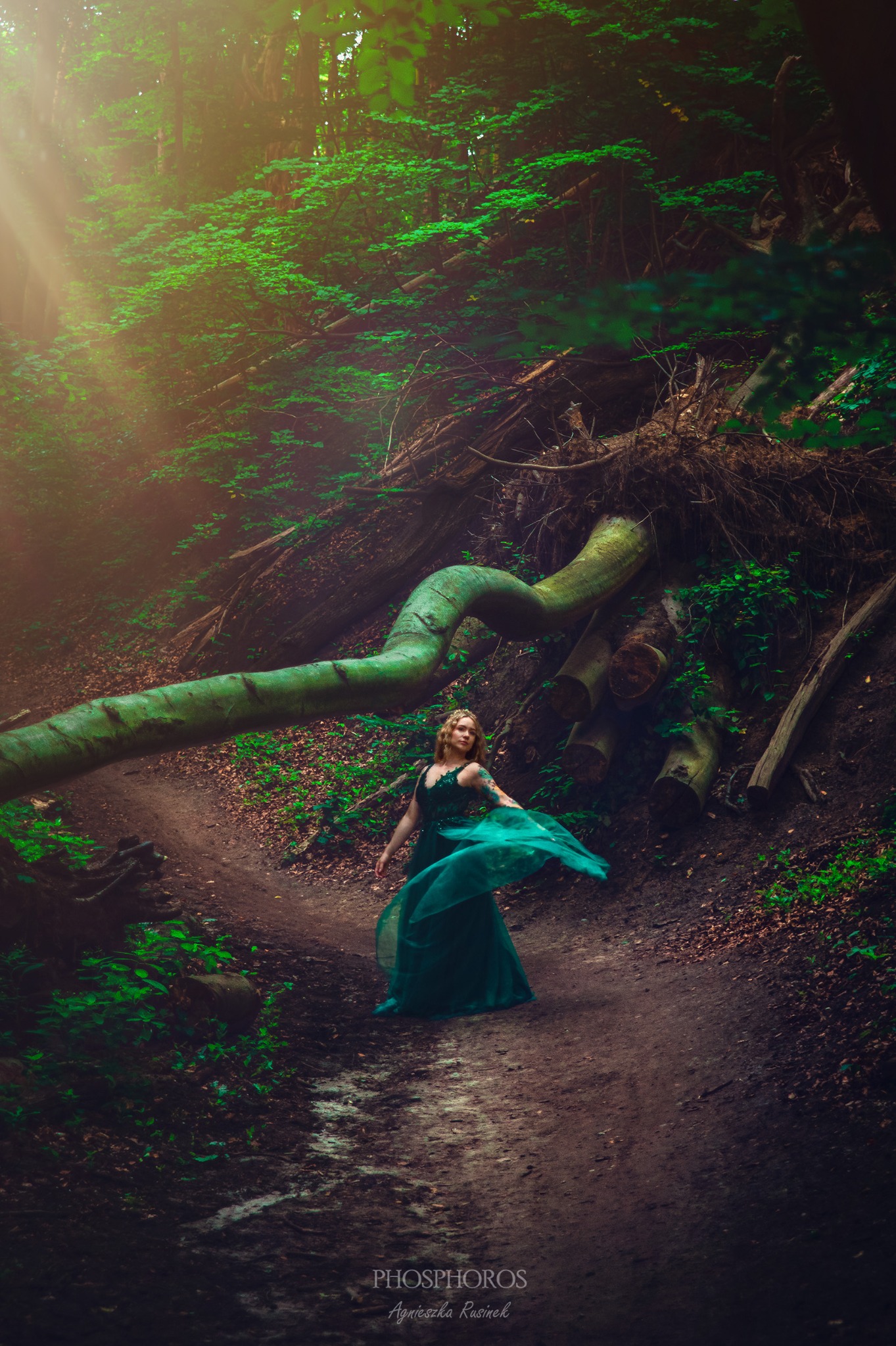 Portret kobiety w zielonej sukni tańczącej w leśnej scenerii. Promienie słońca przebijają się przez korony drzew, oświetlając ścieżkę i podkreślając magiczny charakter zdjęcia.