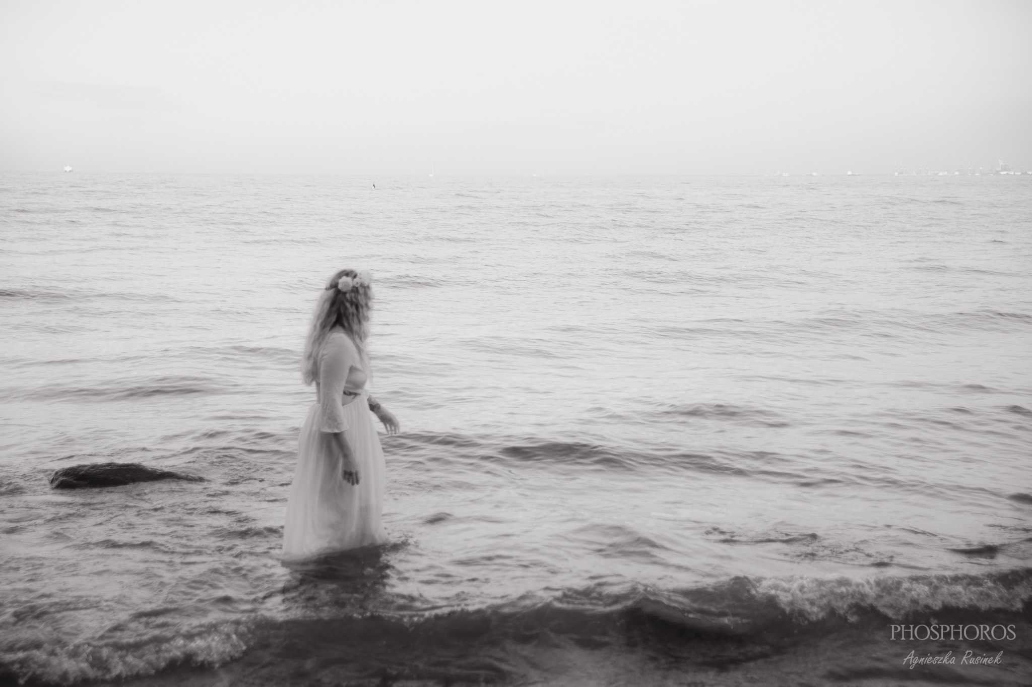 Czarno-białe zdjęcie kobiety w białej sukience i wianku, stojącej w wodzie. Delikatne fale, odległy brzeg. Fotografia plenerowa w Grudziądzu.