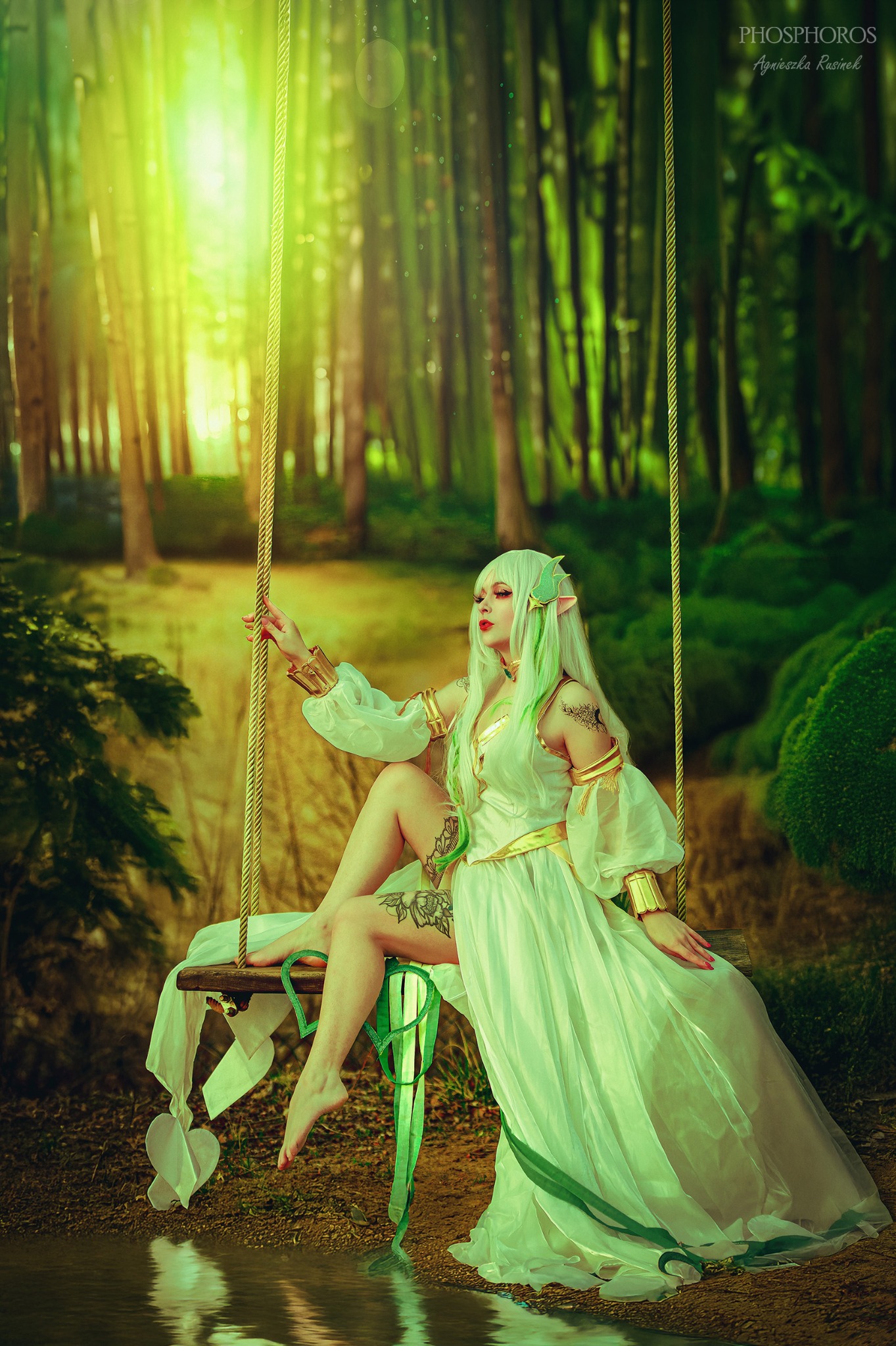 Kobieta w elfickim stroju siedzi na huśtawce w lesie, promienie słońca przebijają przez drzewa, odbijając się w wodzie. Uwagę przykuwa mistyczny klimat i bajkowy charakter sesji.