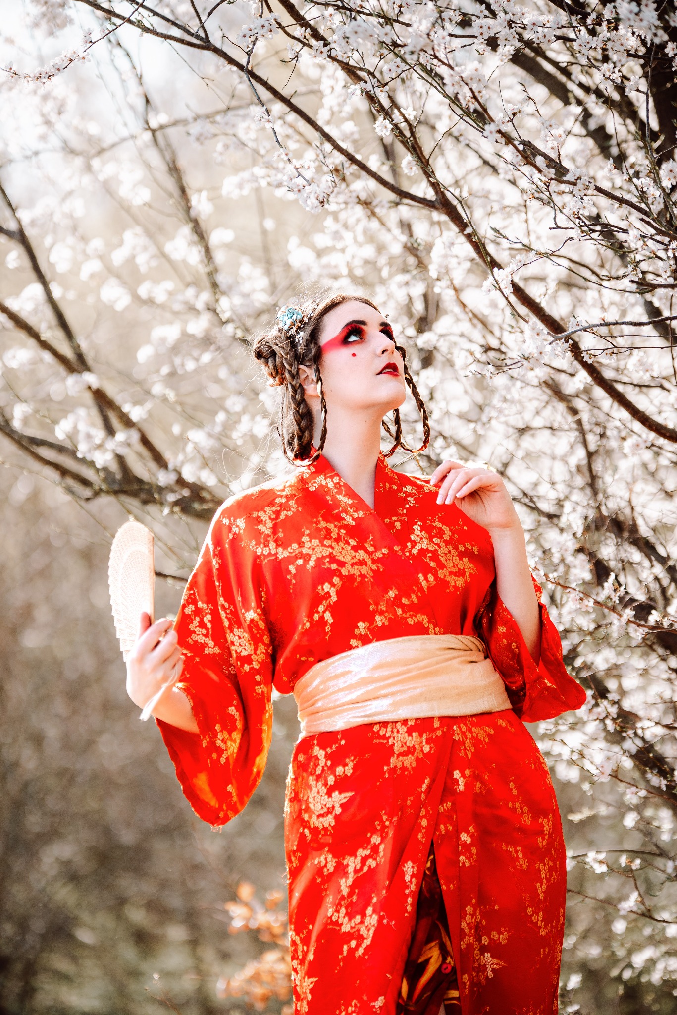 Kobieta w czerwonym kimonie z wachlarzem pozuje na tle kwitnącej wiśni. Uwagę zwraca makijaż z czerwonymi akcentami. Nietypowa sesja w plenerze, artystyczne ujęcie.