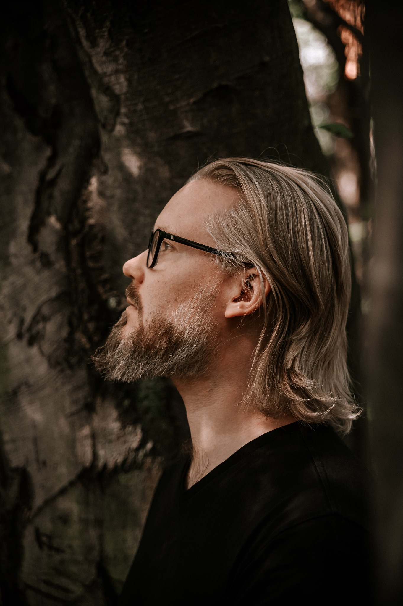 Portret mężczyzny z brodą i okularami, profil, na tle kory drzewa. Stonowana kolorystyka, naturalne światło, artystyczne ujęcie, skupienie na detalu twarzy.