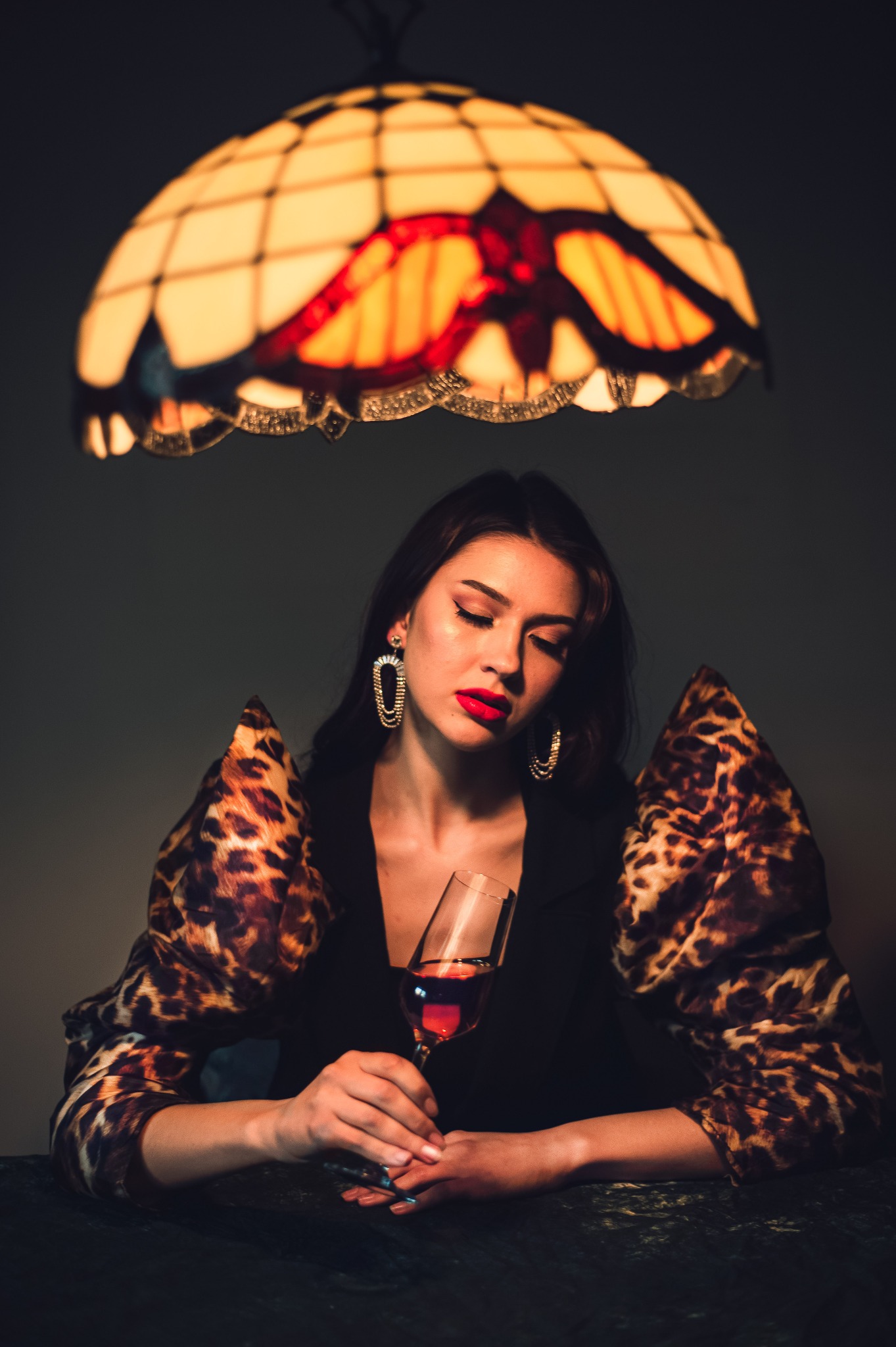 Stylowa sesja: Kobieta w bluzce w panterkę, z czerwoną szminką i drinkiem w dłoni, pod ciepłym światłem lampy Tiffany. Elegancka biżuteria, stonowane tło.