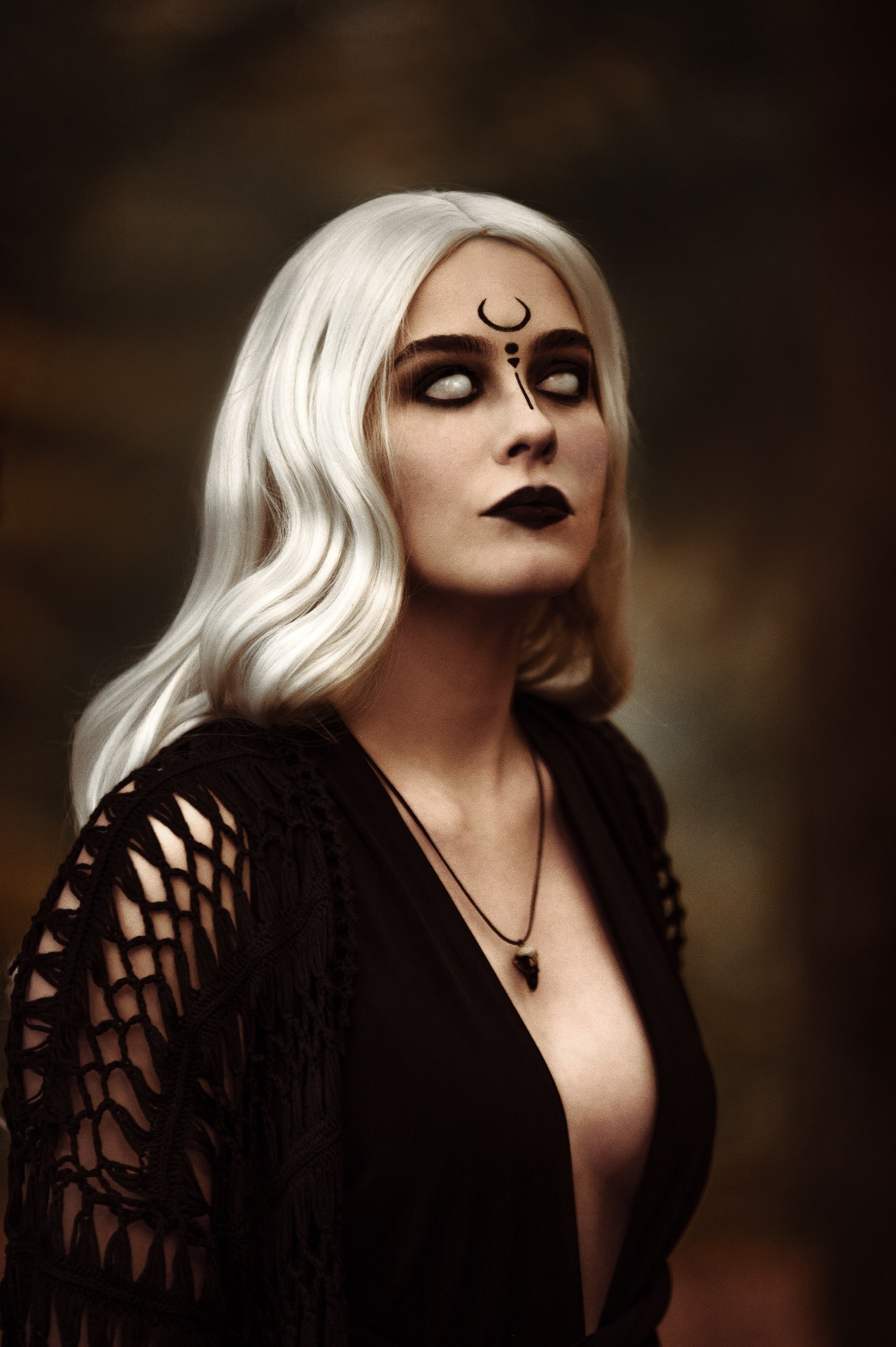 Portret kobiety z bladą cerą, białymi włosami i ciemnym makijażem oraz symbolem na czole, spoglądającej w górę, na rozmytym, ciemnym tle.