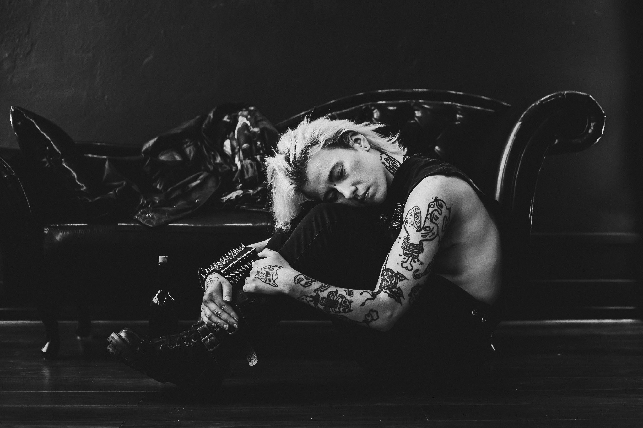 Czarno-białe zdjęcie wytatuowanego mężczyzny z blond włosami, siedzącego na podłodze obok skórzanej kanapy. Wyrazisty styl, ciemne tło, ostre światło.