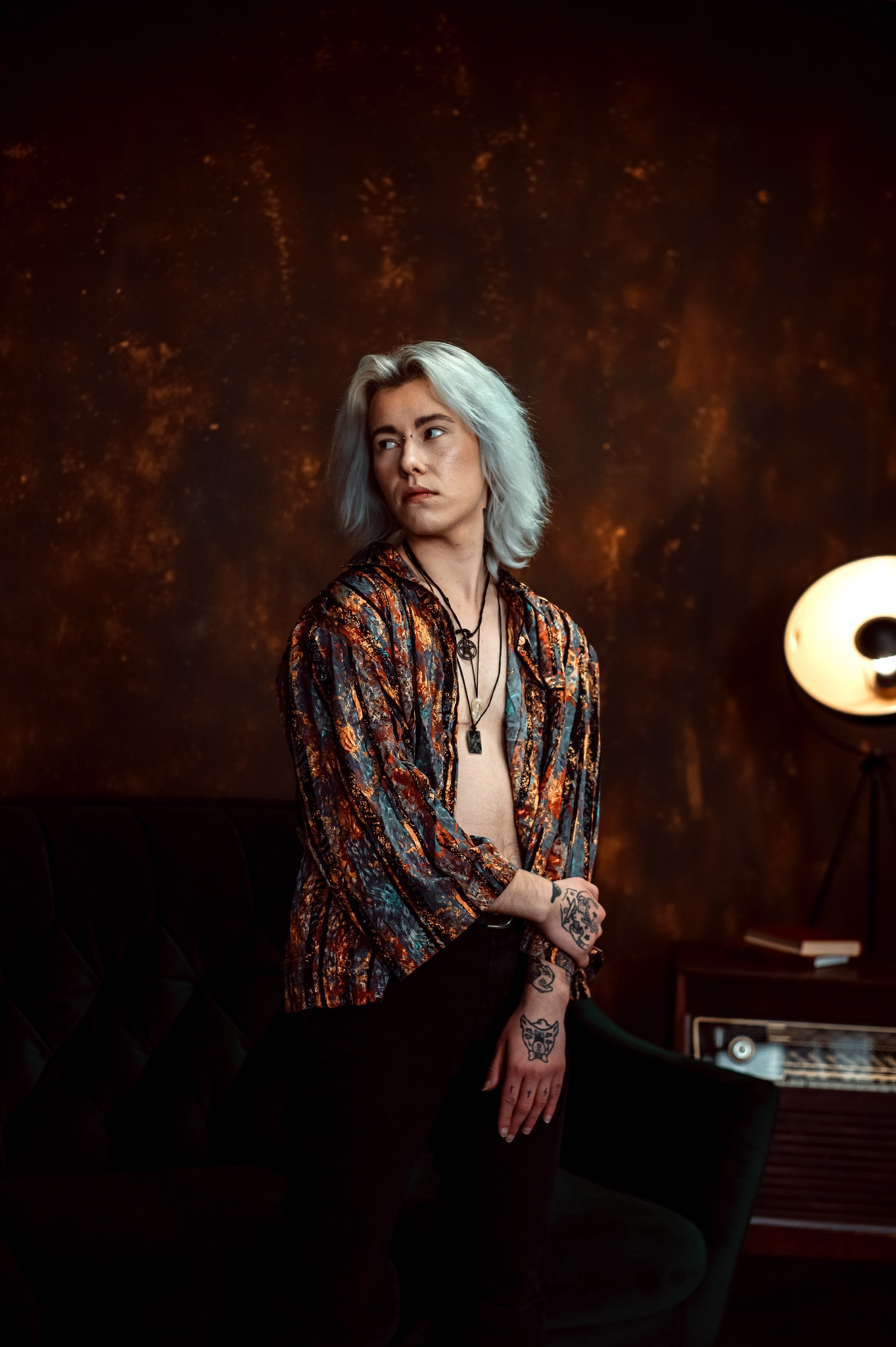 Osoba z platynowymi włosami i tatuażami na dłoniach, w otwartej, wzorzystej koszuli, siedzi na ciemnozielonej sofie na tle brązowej ściany. Obok stoi lampa i radio.