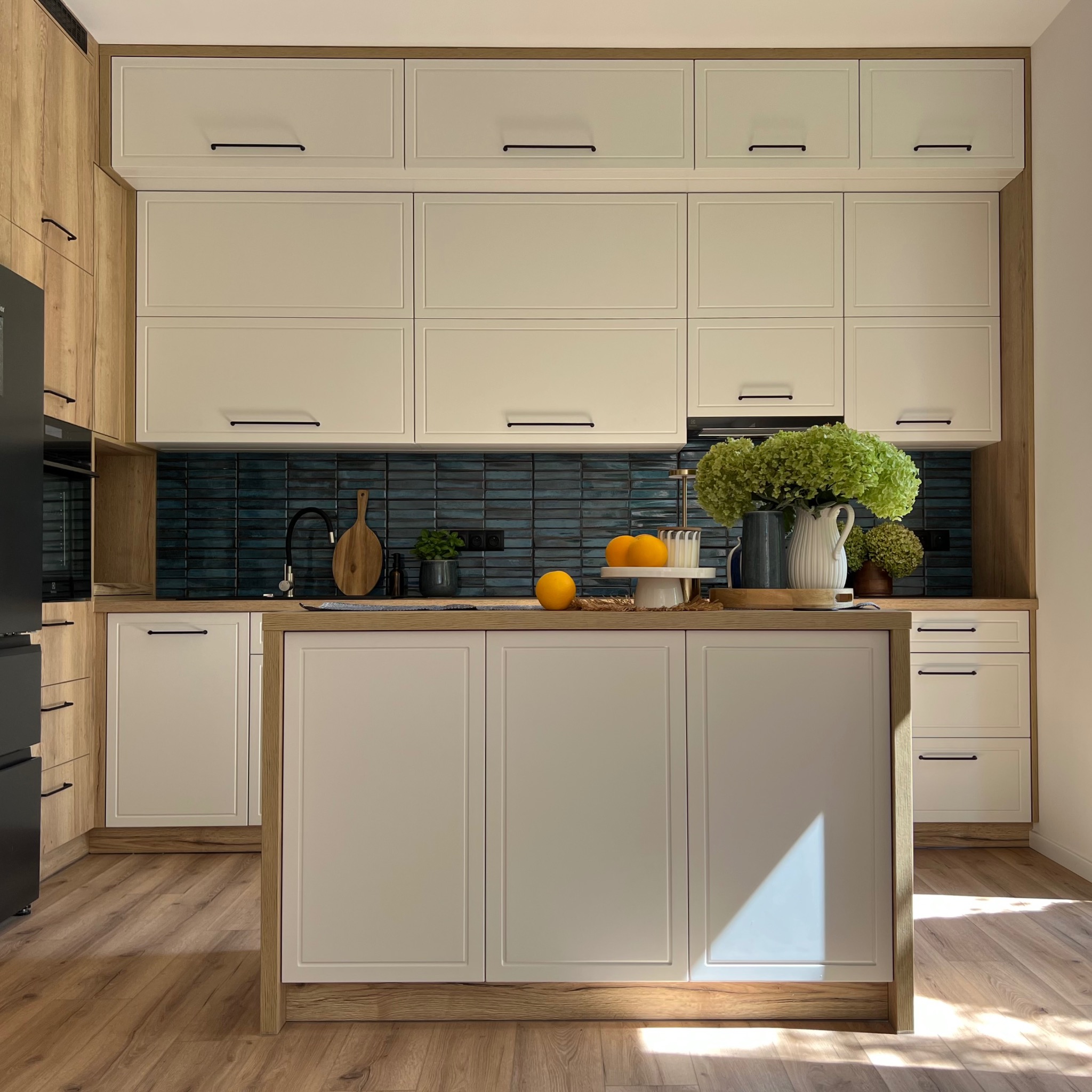 Kuchnia w stylu rustykalnym z wyspą, białe fronty szafek i drewniane akcenty. Na blacie dekoracje: owoce, kwiaty w wazonie, deska do krojenia, ciemna glazura na ścianie.