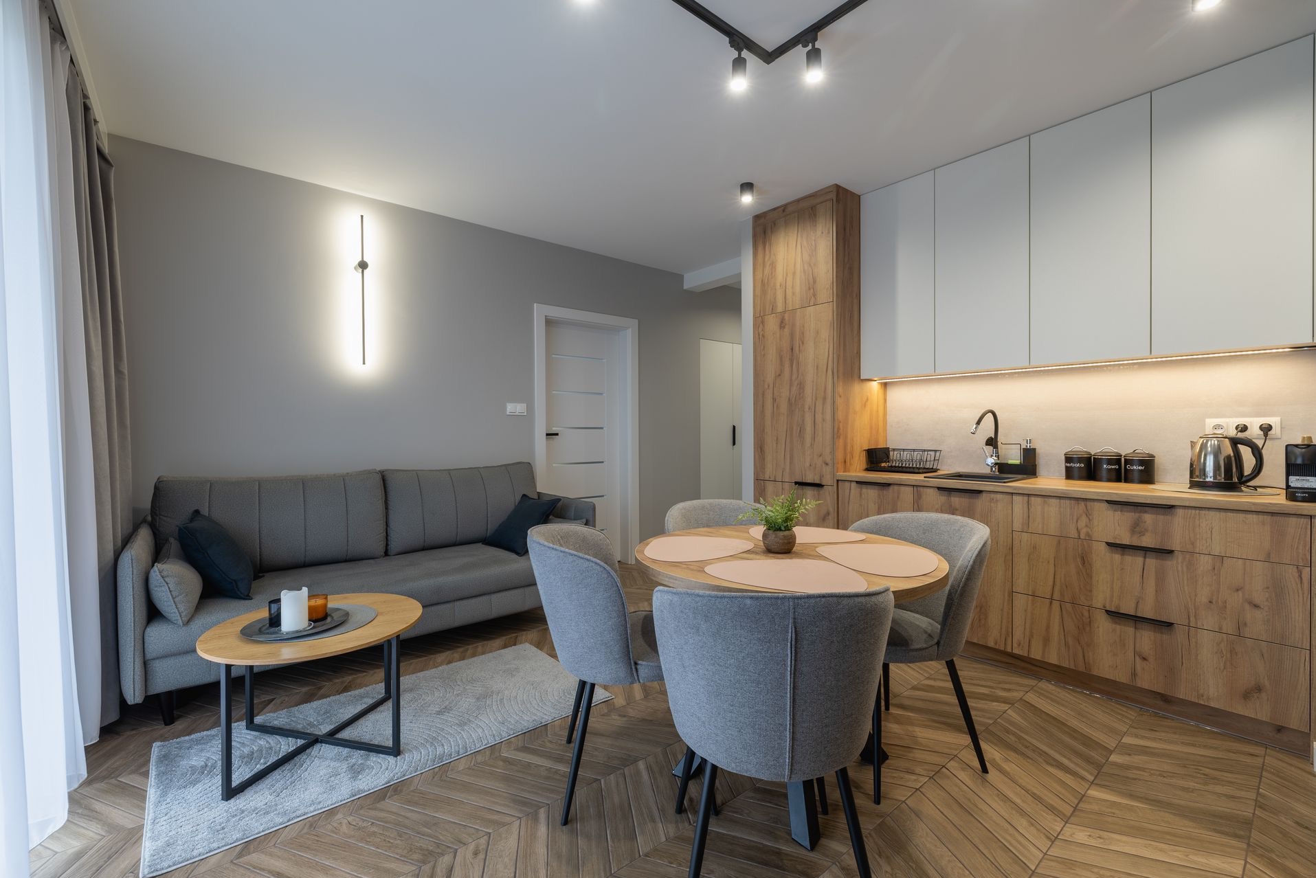 Nowoczesne wnętrze: szara sofa, okrągły stół jadalny z czterema krzesłami, drewniana podłoga w jodełkę i zabudowa kuchenna w odcieniach drewna i bieli.