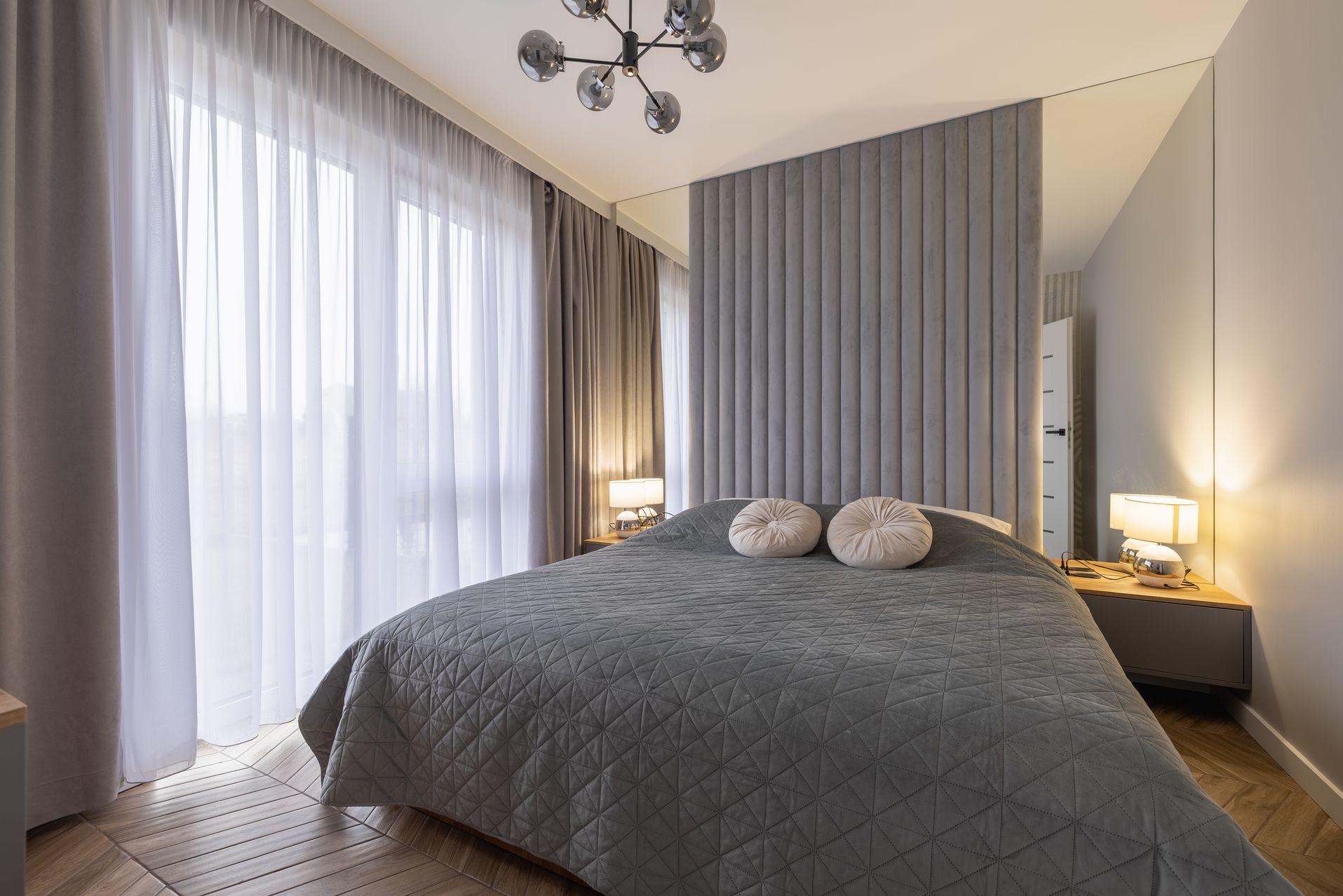 Nowoczesna sypialnia z szarą tapicerowaną ścianą, lustrzanymi panelami i designerską lampą. Uwagę przykuwają okrągłe poduszki na łóżku i parkiet ułożony w jodełkę.