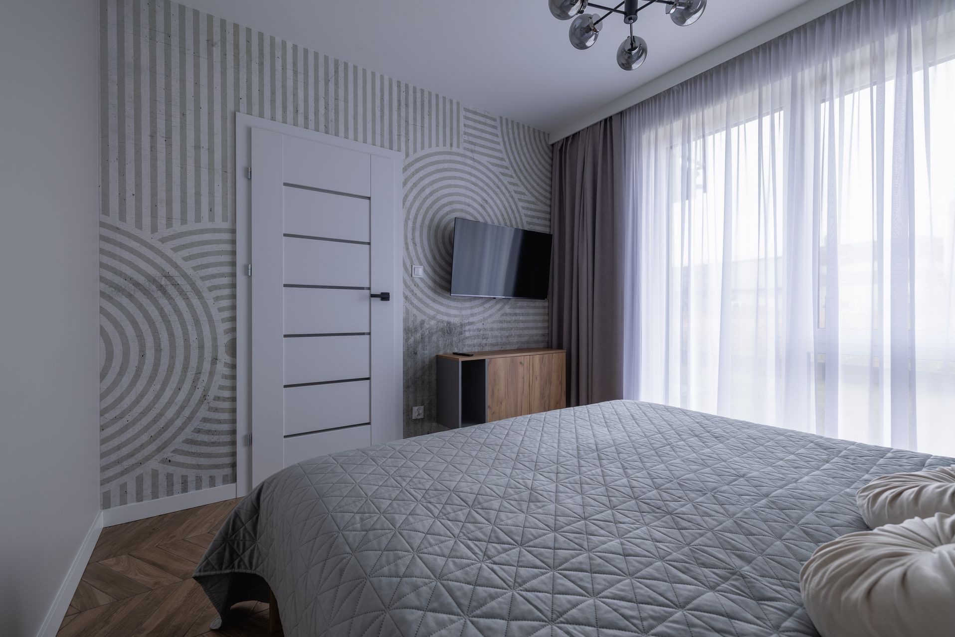Nowoczesna sypialnia z geometrycznym wzorem na ścianie, telewizorem i łóżkiem z szarą narzutą. Jasne zasłony wpuszczają światło dzienne. Widoczny fragment parkietu.
