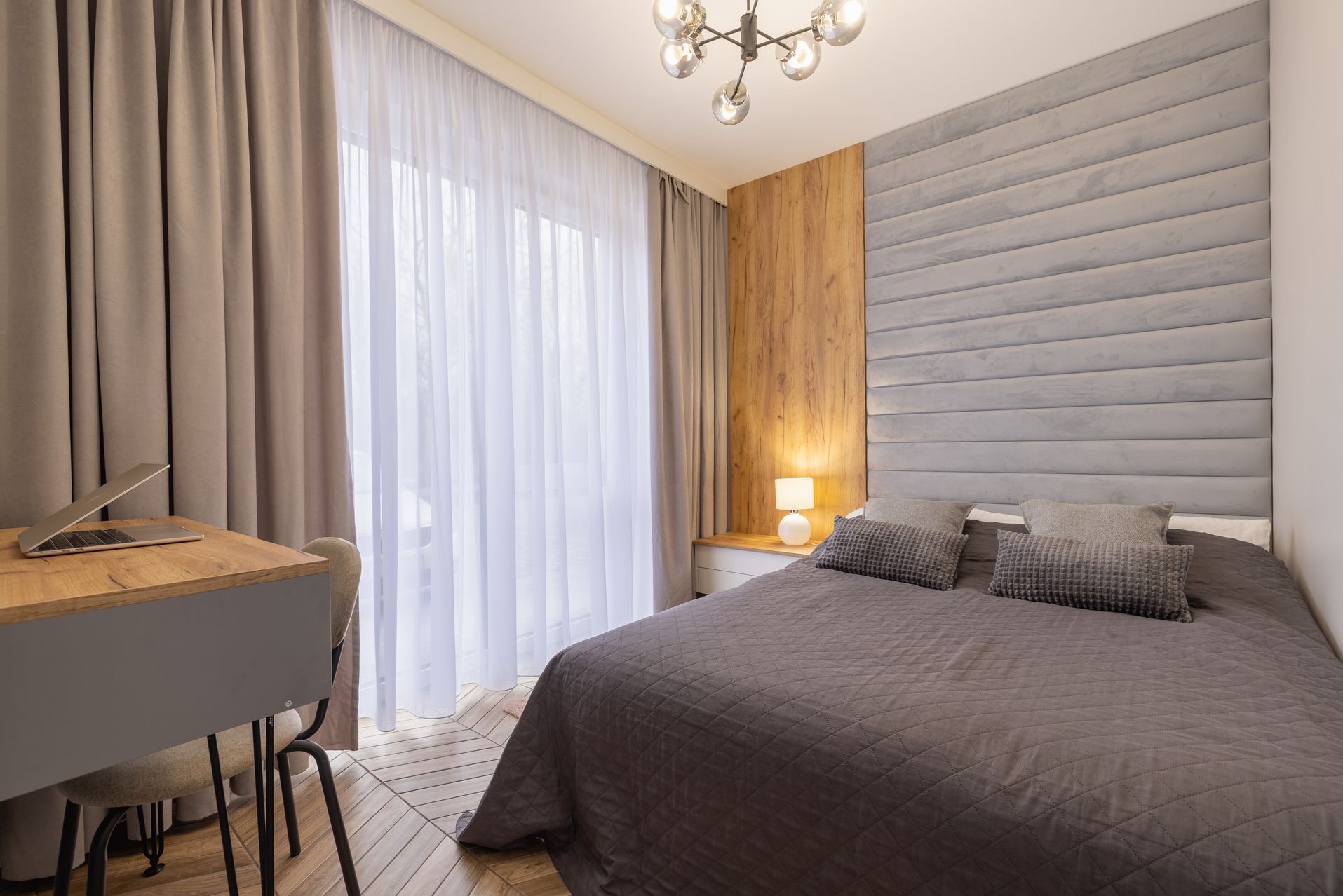 Nowoczesna sypialnia z szarym łóżkiem, drewnianym panelem i biurkiem z laptopem. Subtelne oświetlenie i jasne zasłony tworzą przytulną atmosferę.