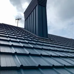 Szary dach z blachy trapezowej z kominem wentylacyjnym i anteną na tle pochmurnego nieba. Widok z dołu, podkreślający regularność wzoru i nowoczesny design.