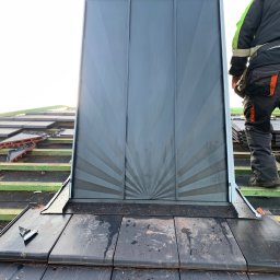 Michalbud - Montaż komina systemowego na dachu pokrytym dachówką ceramiczną. Widoczny fragment dachu, komin z blachy w kolorze grafitowym i fragment sylwetki dekarza.