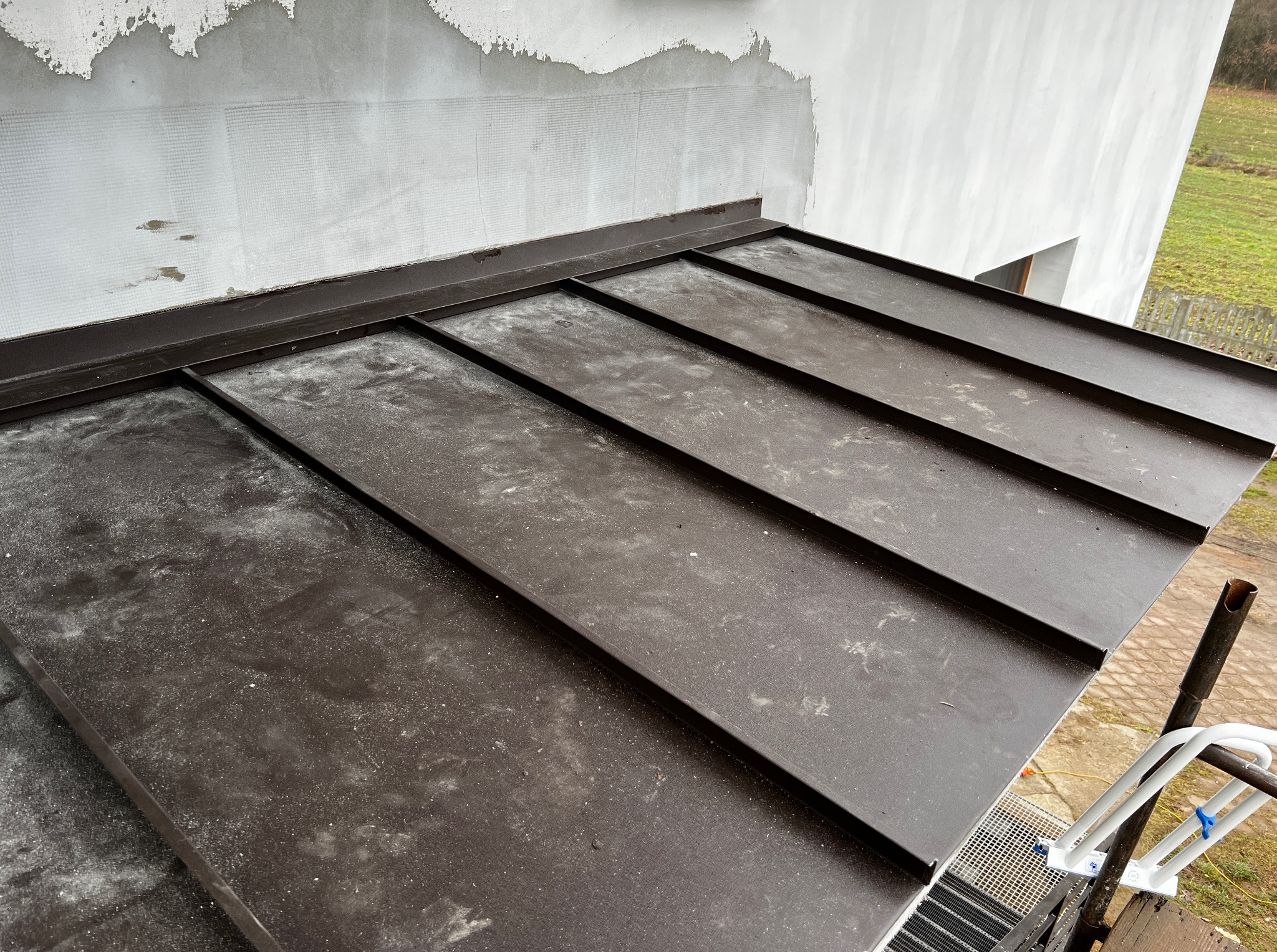 Nowoczesne, brązowe krycie dachu z blachy na budynku w trakcie wykończenia elewacji. Widoczne detale obróbek blacharskich i fragment rusztowania.