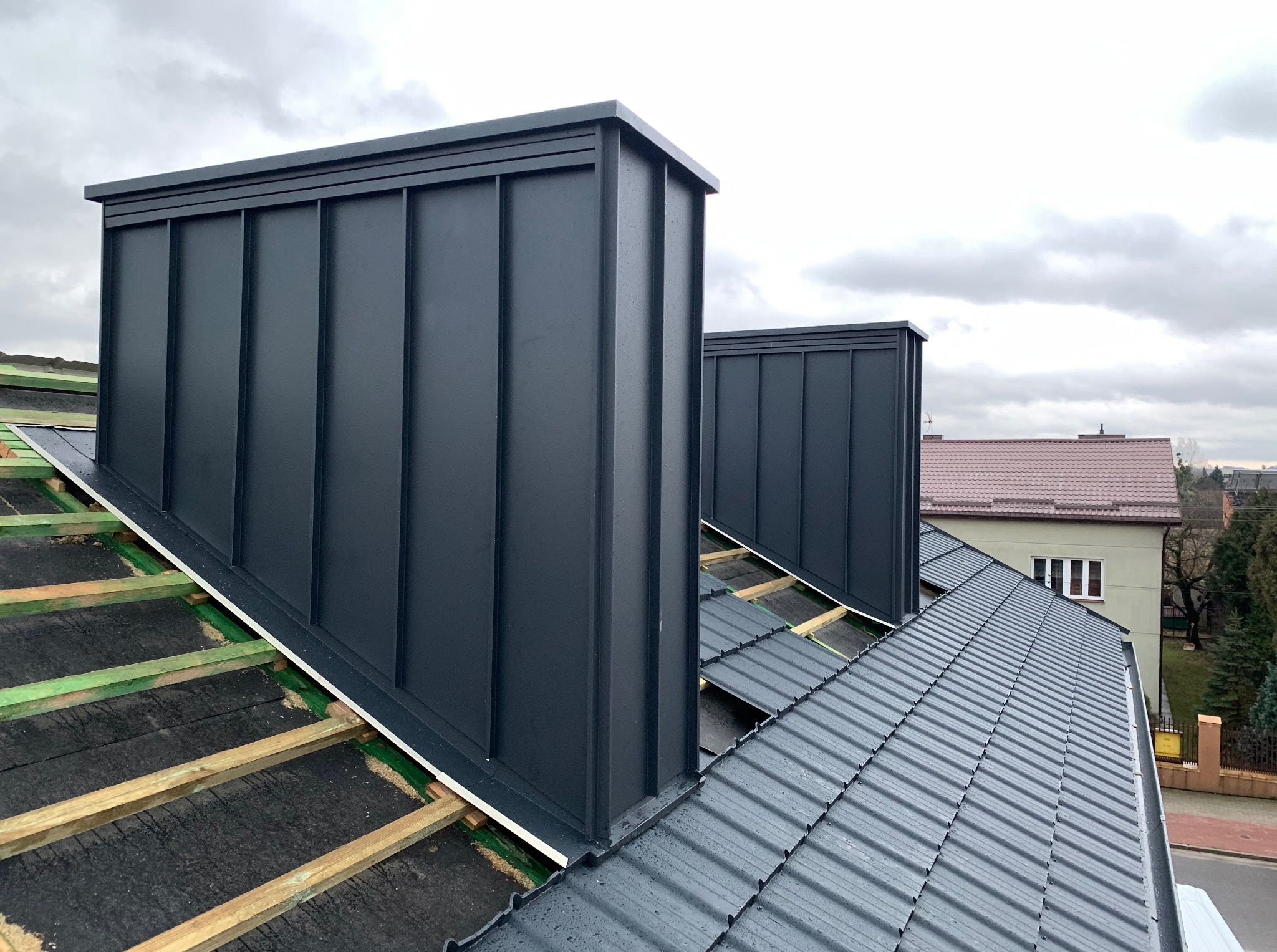 Szary dach z blachy z dwoma kominami w nowoczesnym stylu. Widoczne drewniane elementy konstrukcji dachu i fragmenty rynny. W tle budynki mieszkalne i pochmurne niebo.