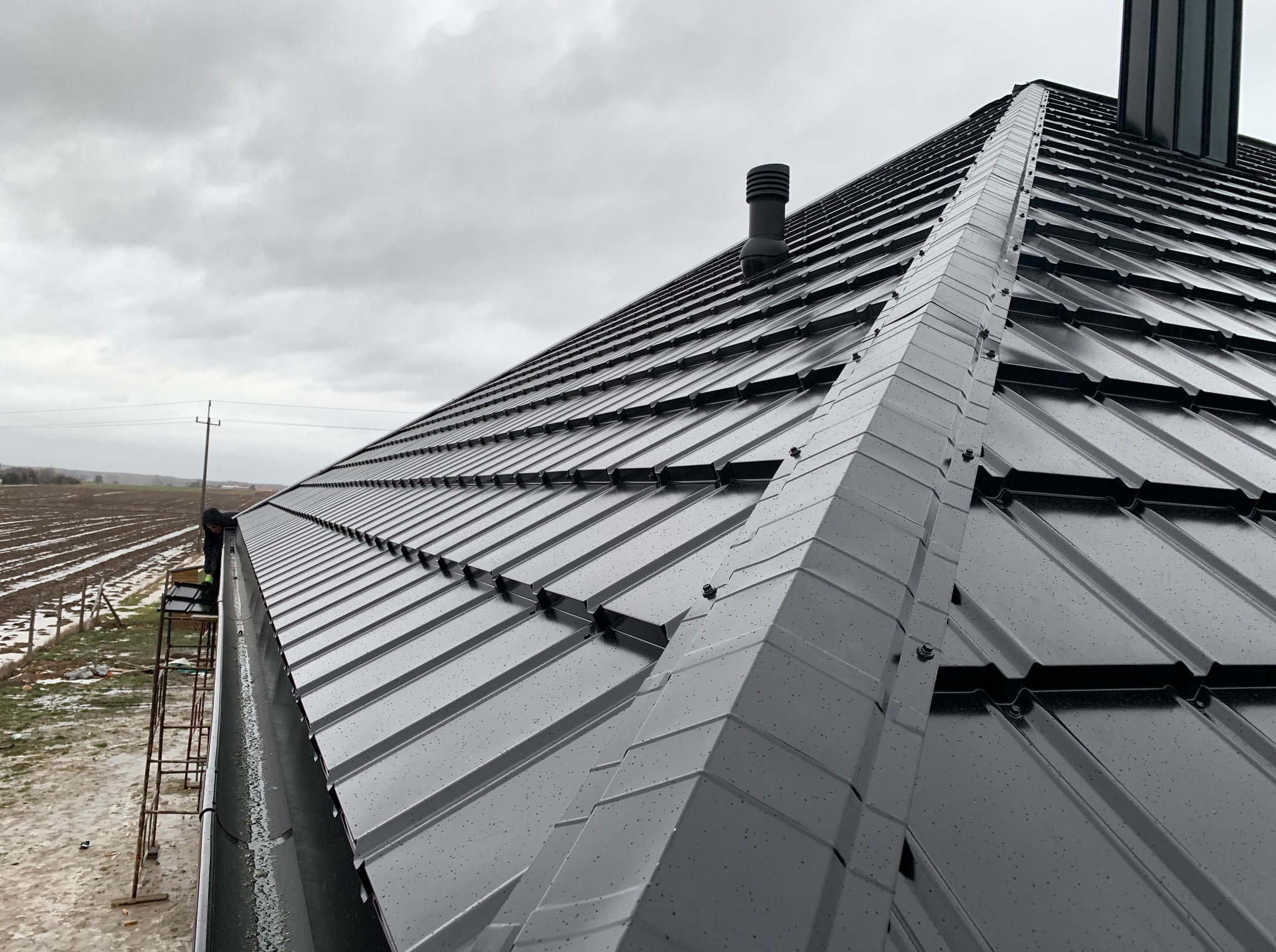 Czarny, metalowy dach z kominem i orynnowaniem w pochmurny dzień. Widoczna drabina i fragment krajobrazu w tle. Ujęcie z dołu, podkreślające geometrię dachu.