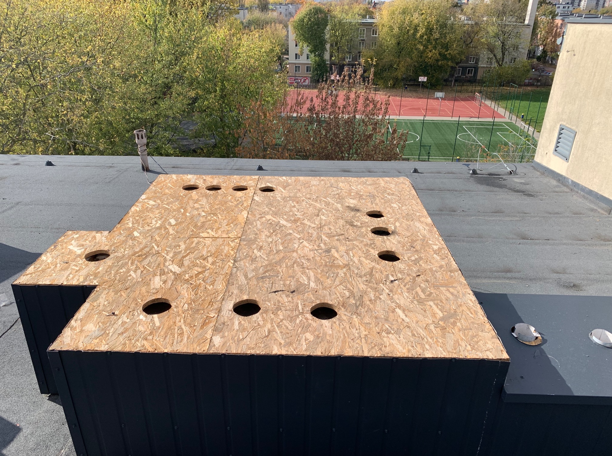 Płyta OSB z wyciętymi otworami na dachu z papy na tle boiska i drzew. Element konstrukcyjny przygotowany do dalszych prac dekarskich.