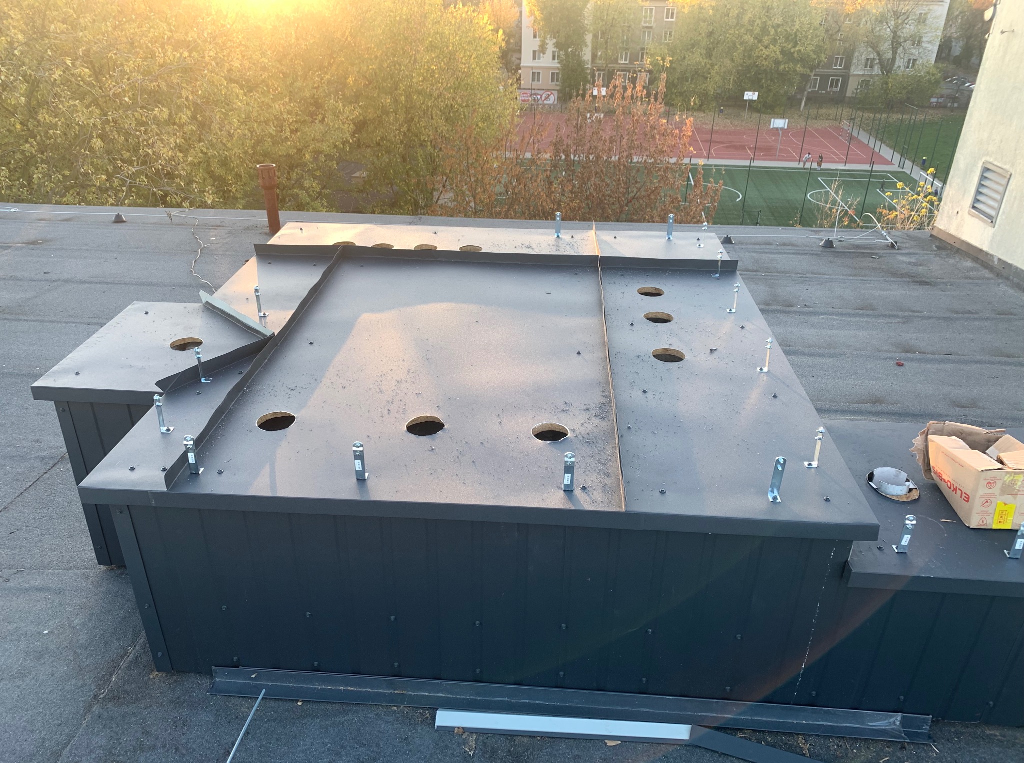 Czarna, metalowa konstrukcja na dachu budynku z otworami i wystającymi śrubami. W tle drzewa i budynki w popołudniowym świetle. Widoczne narzędzia i elementy montażowe.