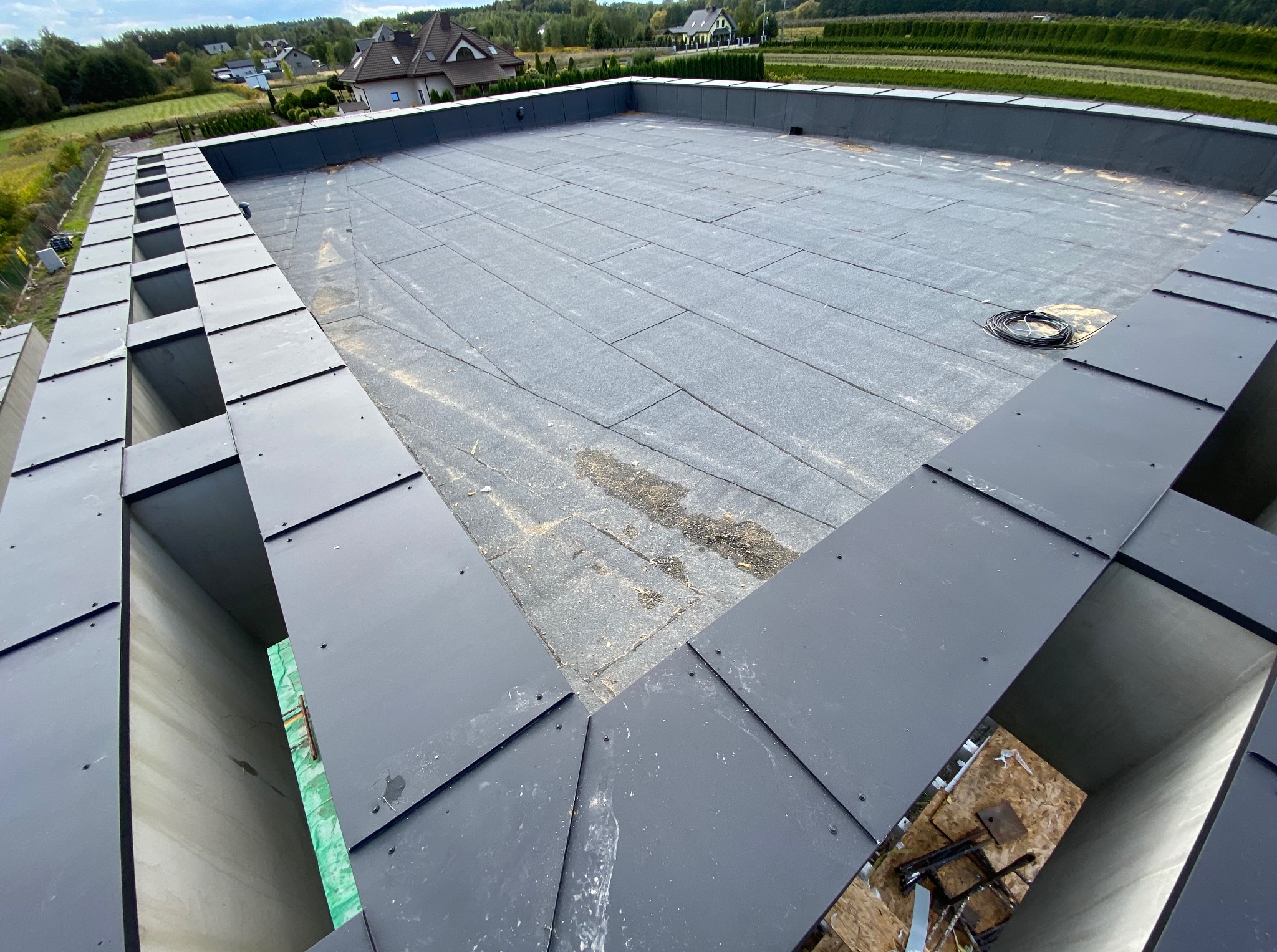 Płaski dach pokryty szarą papą z widocznymi obróbkami blacharskimi w kolorze grafitowym. Widok z góry na ukończony dach. W tle zabudowa jednorodzinna i zieleń.