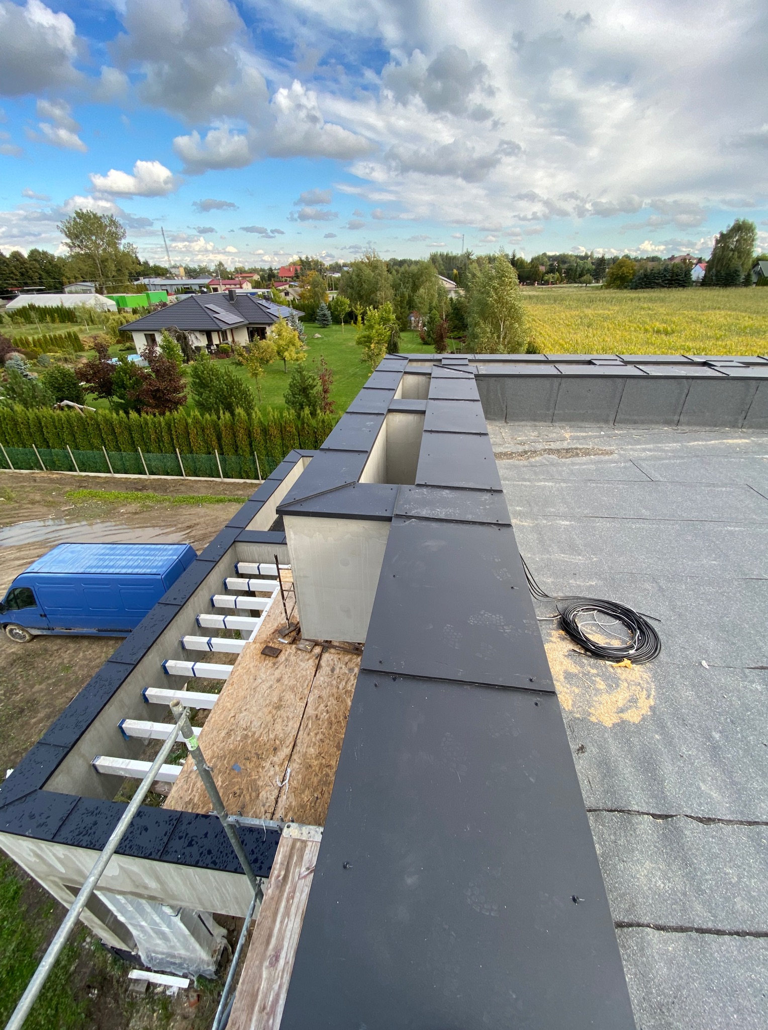 Czarny dach płaski z obróbkami blacharskimi w trakcie montażu, widok z góry. Na dachu leżą kable, w tle zielony krajobraz i błękitny samochód dostawczy.