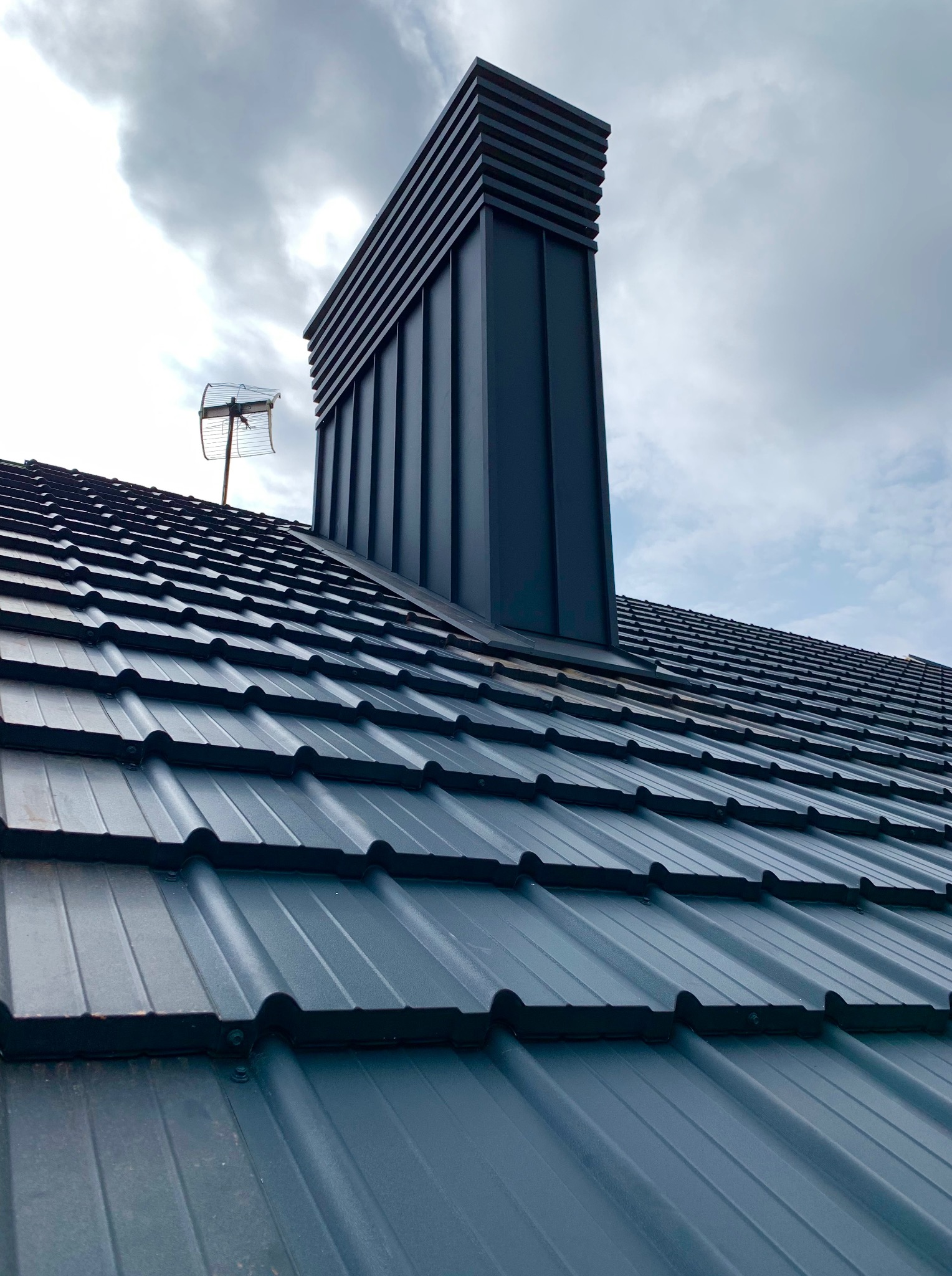 Szary dach z blachy trapezowej z kominem wentylacyjnym i anteną na tle pochmurnego nieba. Widok z dołu, podkreślający regularność wzoru i nowoczesny design.
