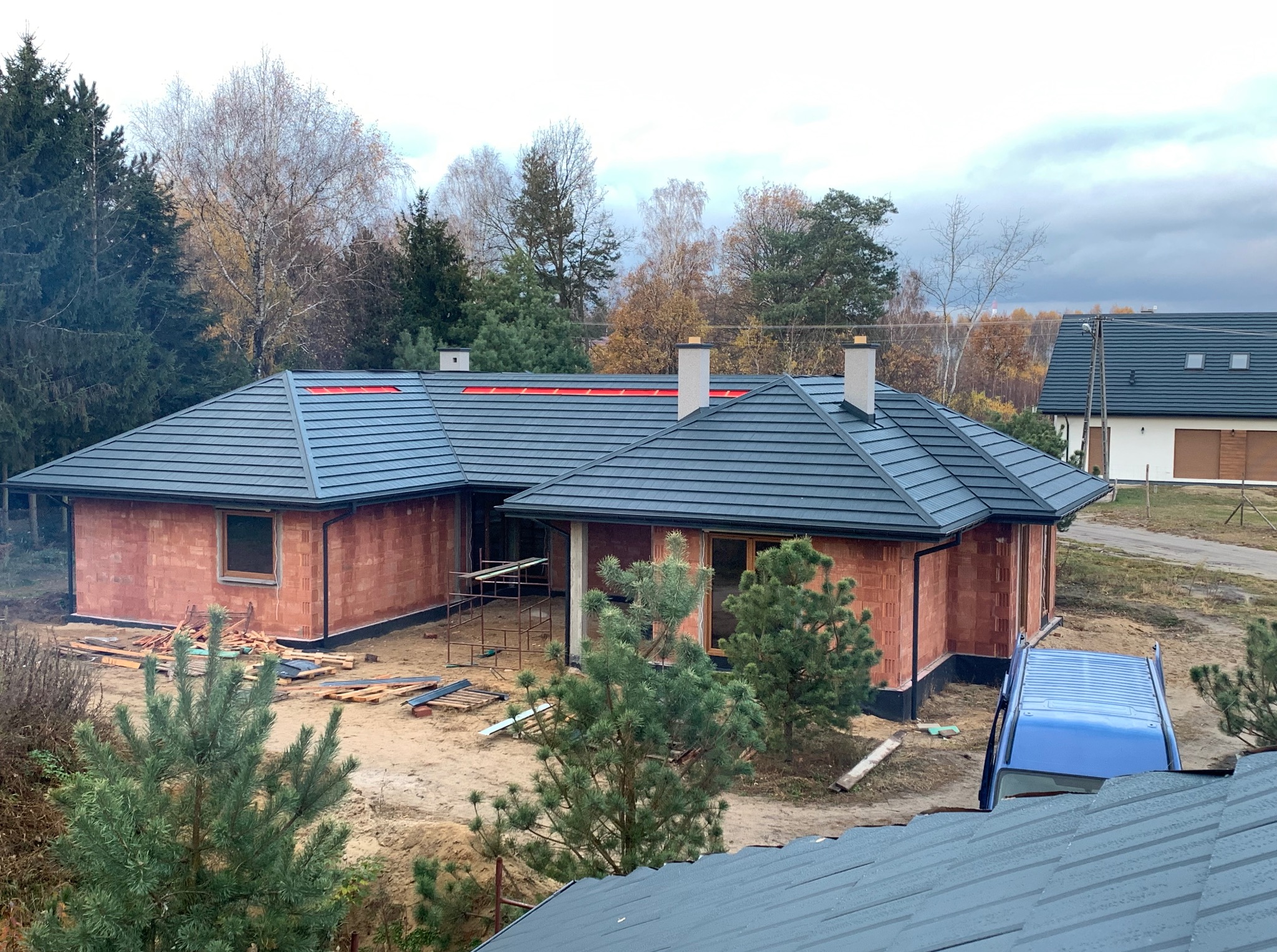 Dom w trakcie budowy z grafitowym dachem z blachy na rąbek stojący i kominami. Widoczny samochód dostawczy i drzewa w tle.