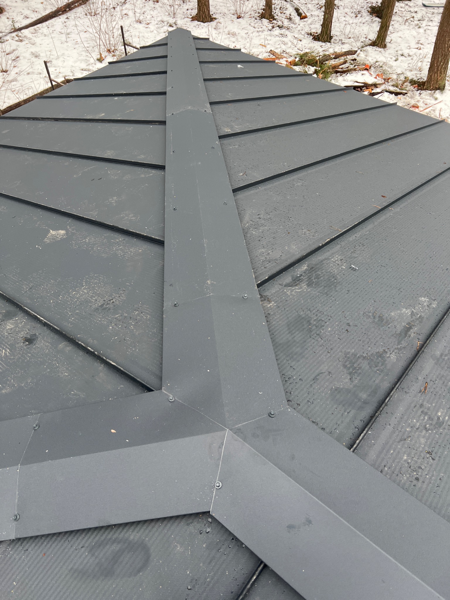 Szary dach z blachy na tle zimowego krajobrazu. Widoczne łączenia blacharskie i śnieg na drzewach w tle. Ujęcie z góry.