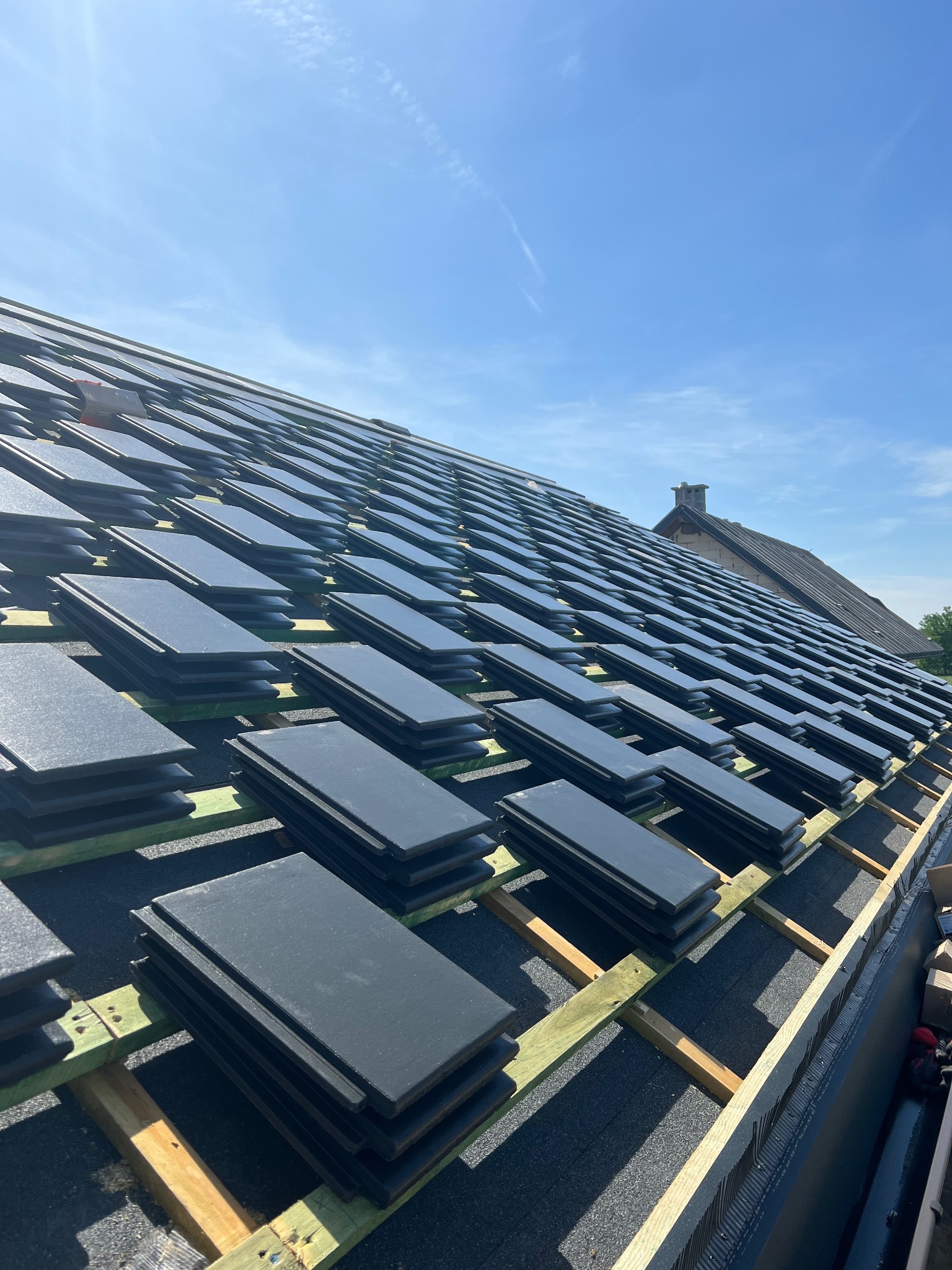 Układanie ciemnych dachówek na drewnianej konstrukcji dachu w słoneczny dzień. Widoczny fragment budynku w tle. Dachówki ułożone w stosy, przygotowane do montażu.