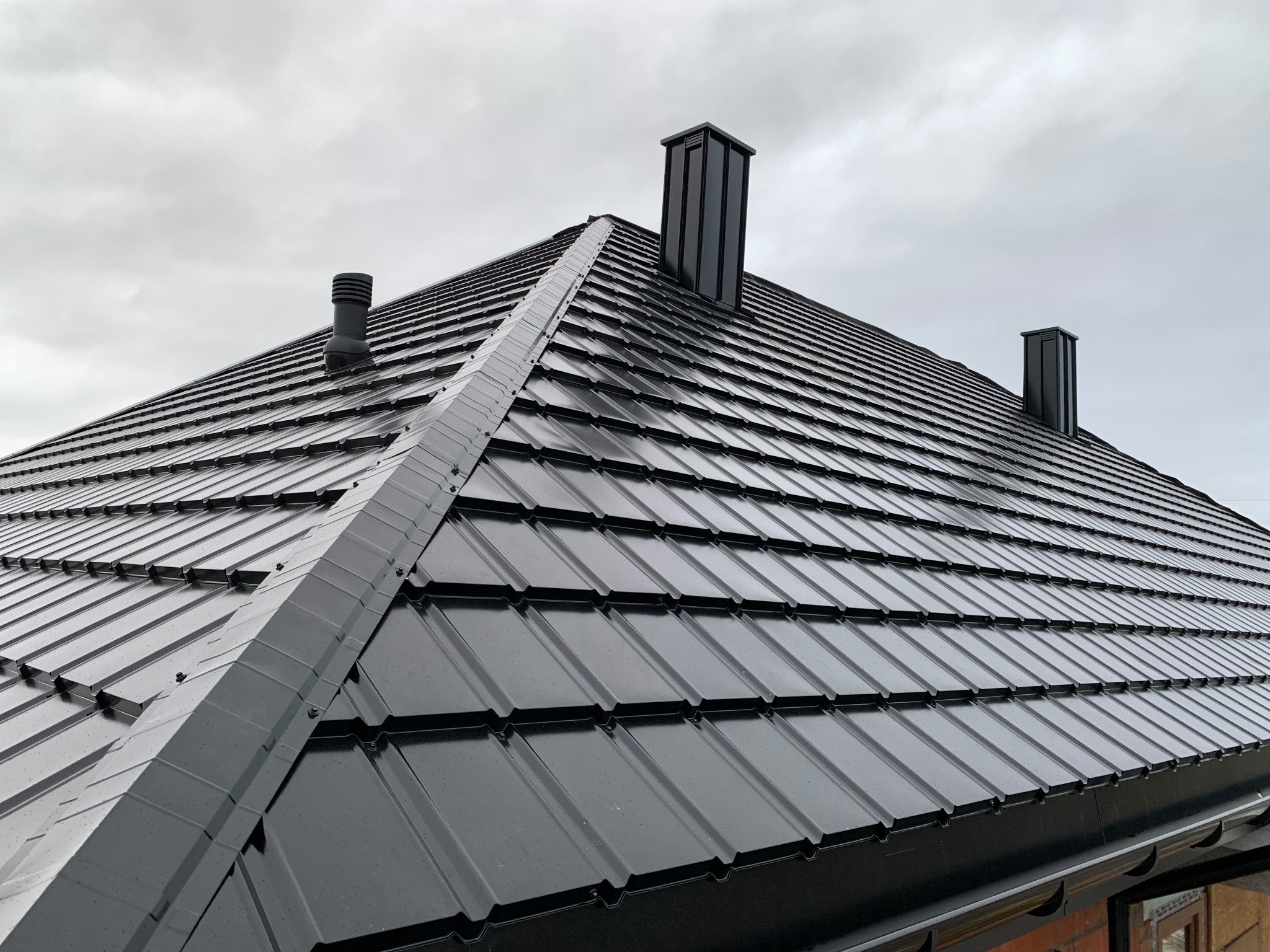 Czarny dach z blachy trapezowej z trzema kominami na tle pochmurnego nieba. Widoczne detale łączeń blachy i obróbek blacharskich.