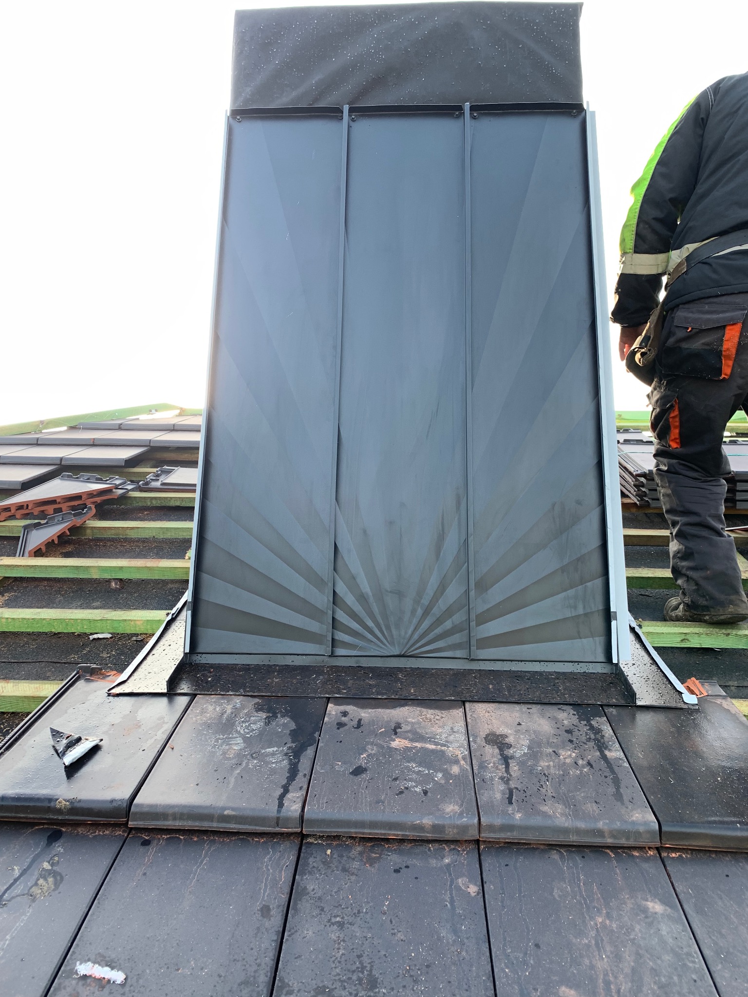 Montaż komina systemowego na dachu pokrytym dachówką ceramiczną. Widoczny fragment dachu, komin z blachy w kolorze grafitowym i fragment sylwetki dekarza.