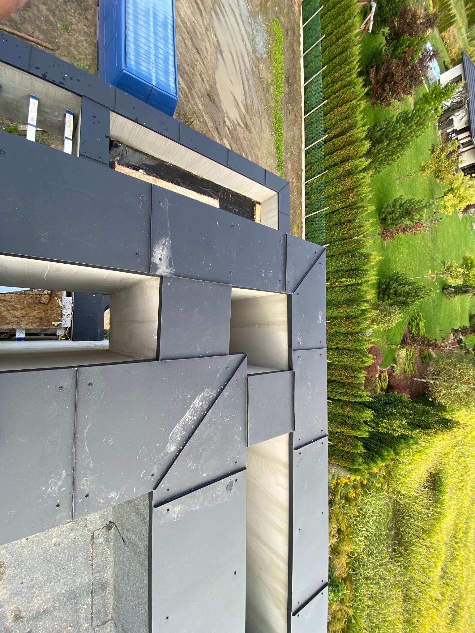 Nowoczesny dach z szarej blachy na betonowej konstrukcji, widok z góry na tle zieleni. Elementy mocowane na nity.